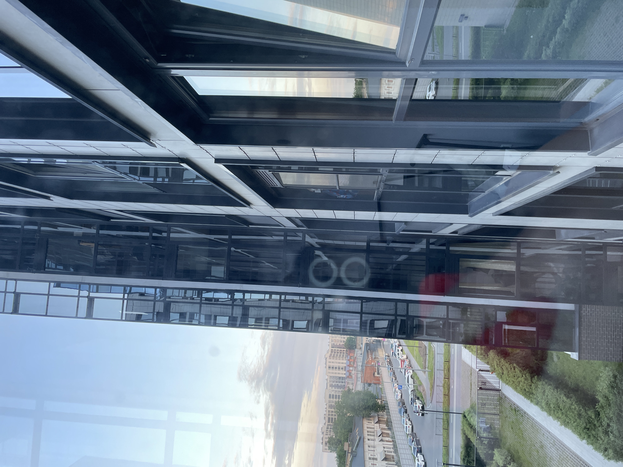 The image shows a modern building with a glass facade, reflecting the sky and surrounding cityscape. In the foreground, the building's dark metal and glass exterior is prominent, with visible window frames and reflections. To the left, a street is visible lined with parked cars and trees, with low-rise buildings in the background. Further in the distance, a large residential complex with light-colored facades is apparent. The sky is partly cloudy, with soft light suggesting it is either early morning or late afternoon. The scene is likely a residential or mixed-use urban environment.