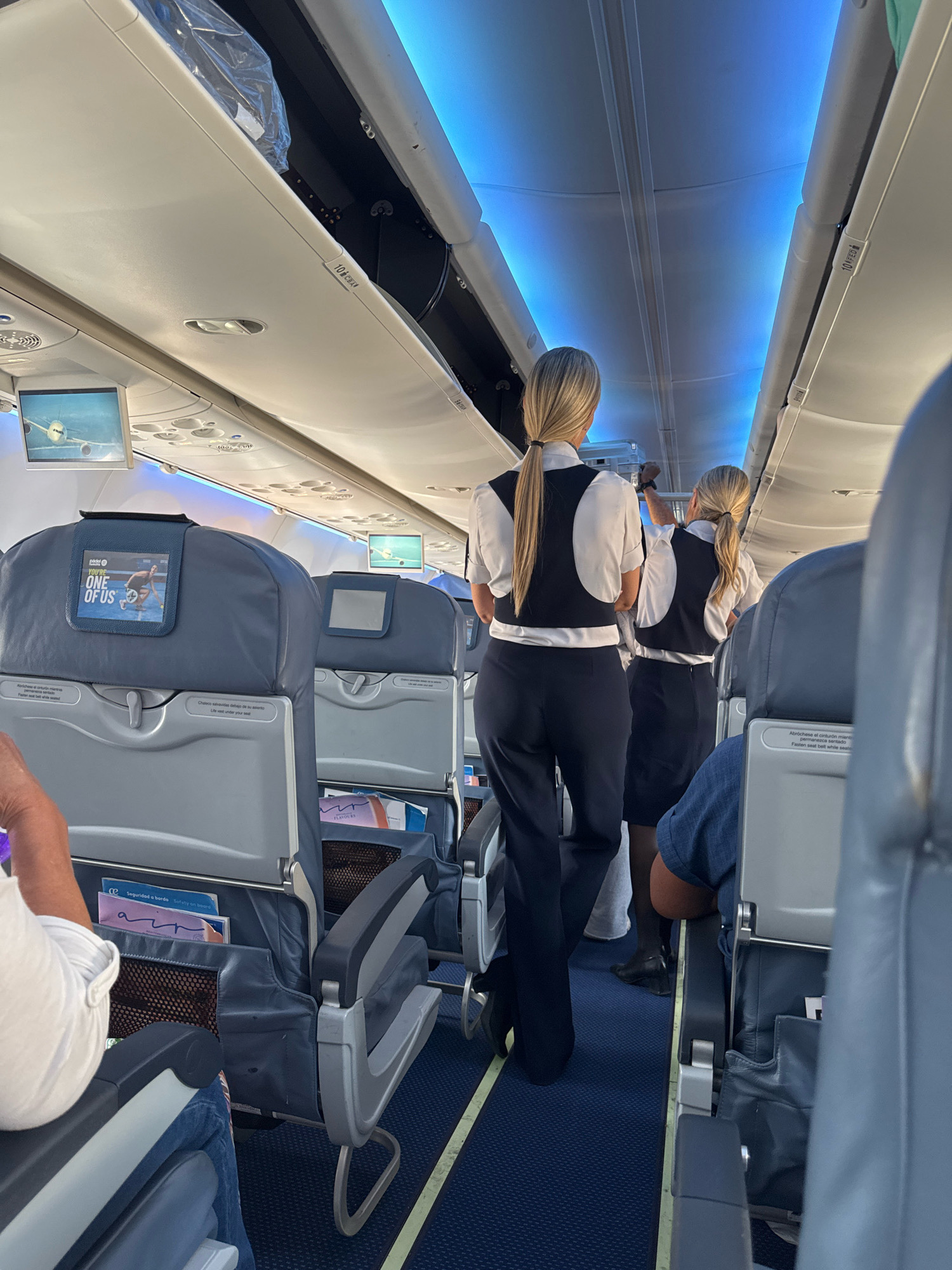 The image depicts the interior of an airplane cabin, viewed from a passenger's perspective. Two flight attendants, dressed in dark vests over white shirts and dark pants/skirts, are walking down the aisle. The cabin features rows of blue upholstered seats, some with individual entertainment screens. Overhead compartments are visible, with blue ambient lighting illuminating the ceiling. The overall environment suggests a commercial flight, likely preparing for departure or during a segment of travel. The mention of Palma de Mallorca, Spain, indicates the potential location of the flight, either originating from, arriving at, or passing through the city.