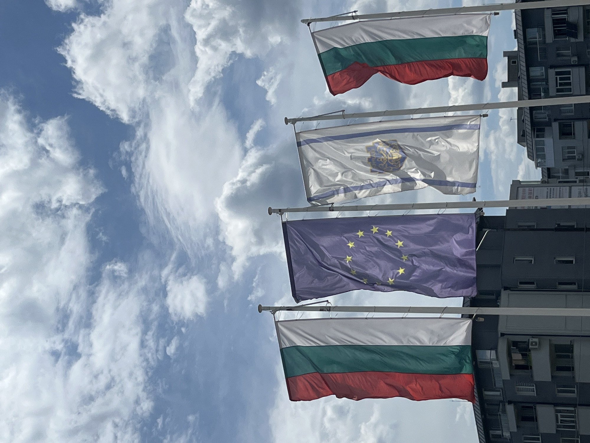The image shows four flags fluttering against a blue sky with scattered clouds, positioned in front of a multi-story building. From top to bottom, the flags are: the Bulgarian flag (white, green, red horizontal stripes), a white flag with a blue border and a golden emblem in the center, the European Union flag (blue with twelve yellow stars), and another Bulgarian flag. The building behind the flags appears to be an apartment complex, a common type of structure in urban areas such as Varna, Bulgaria. The scene suggests a public or institutional setting where these flags are displayed.