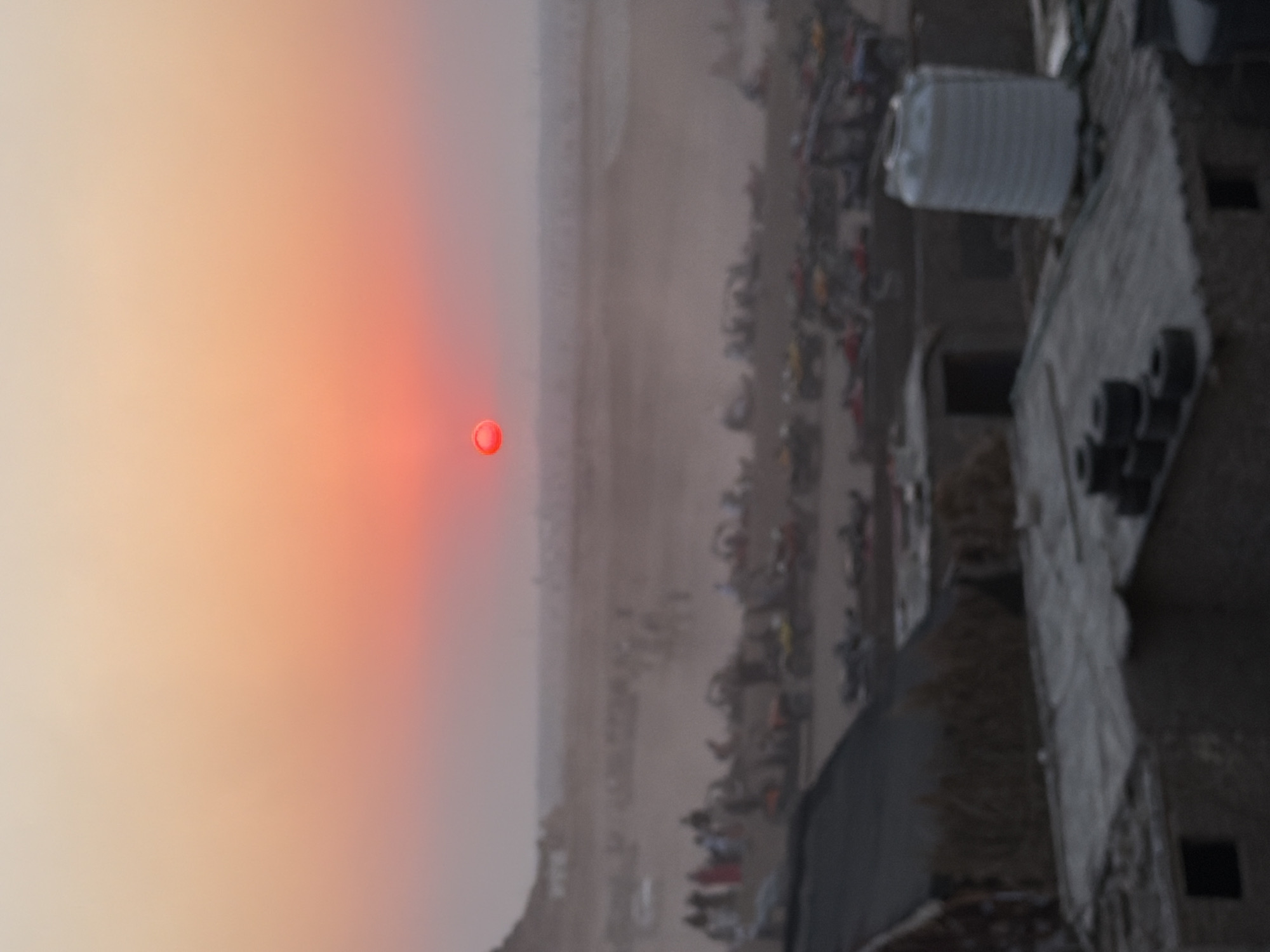 The image depicts a desert landscape at dusk, likely in or around Sharm El Sheikh, Egypt. A large, bright orange sun hangs low in the hazy, gradient sky. In the middle ground, a procession of off-road vehicles, possibly ATVs or dune buggies, are gathered. Many people are visible, some standing near the vehicles and others appearing to be walking in a line. The terrain is sandy and appears to be a popular spot for recreational activities. In the foreground, the roofs of several low-rise buildings are visible, some with corrugated metal roofing. A large, white cylindrical water tank stands on one of the rooftops. The overall impression is one of an outdoor recreational area at sunset.