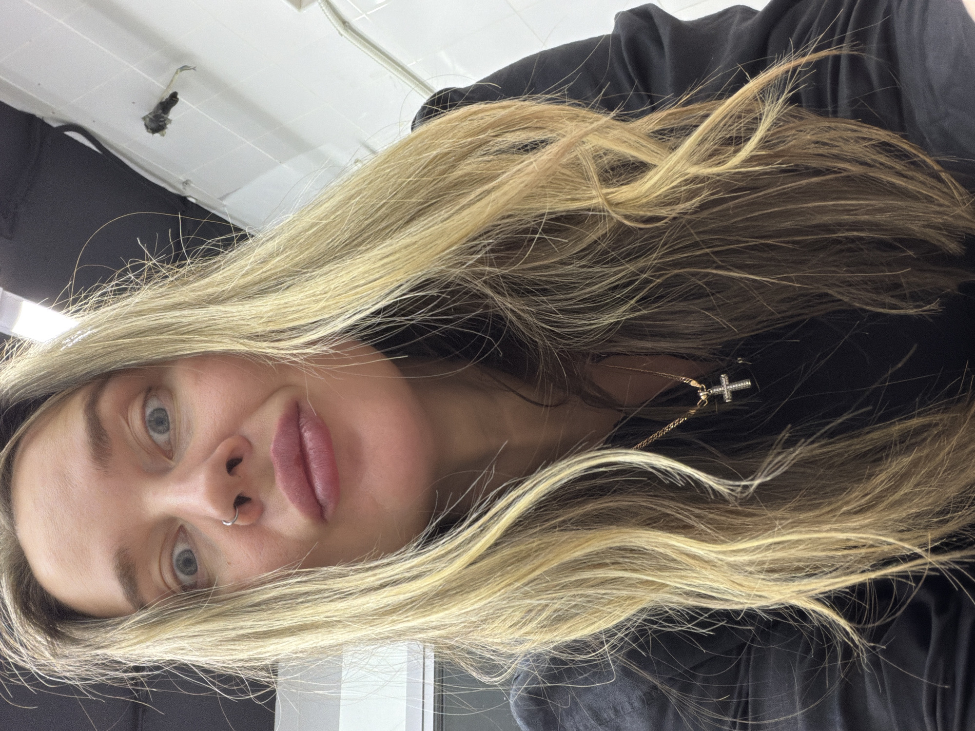 A woman with blonde hair and a nose ring is positioned in the center of the frame. She is wearing a black top and a necklace with a cross pendant. The background includes a ceiling and what appears to be a black curtain or clothing item. The location is not discernible from the image and cannot be placed in Moscow, Russia.