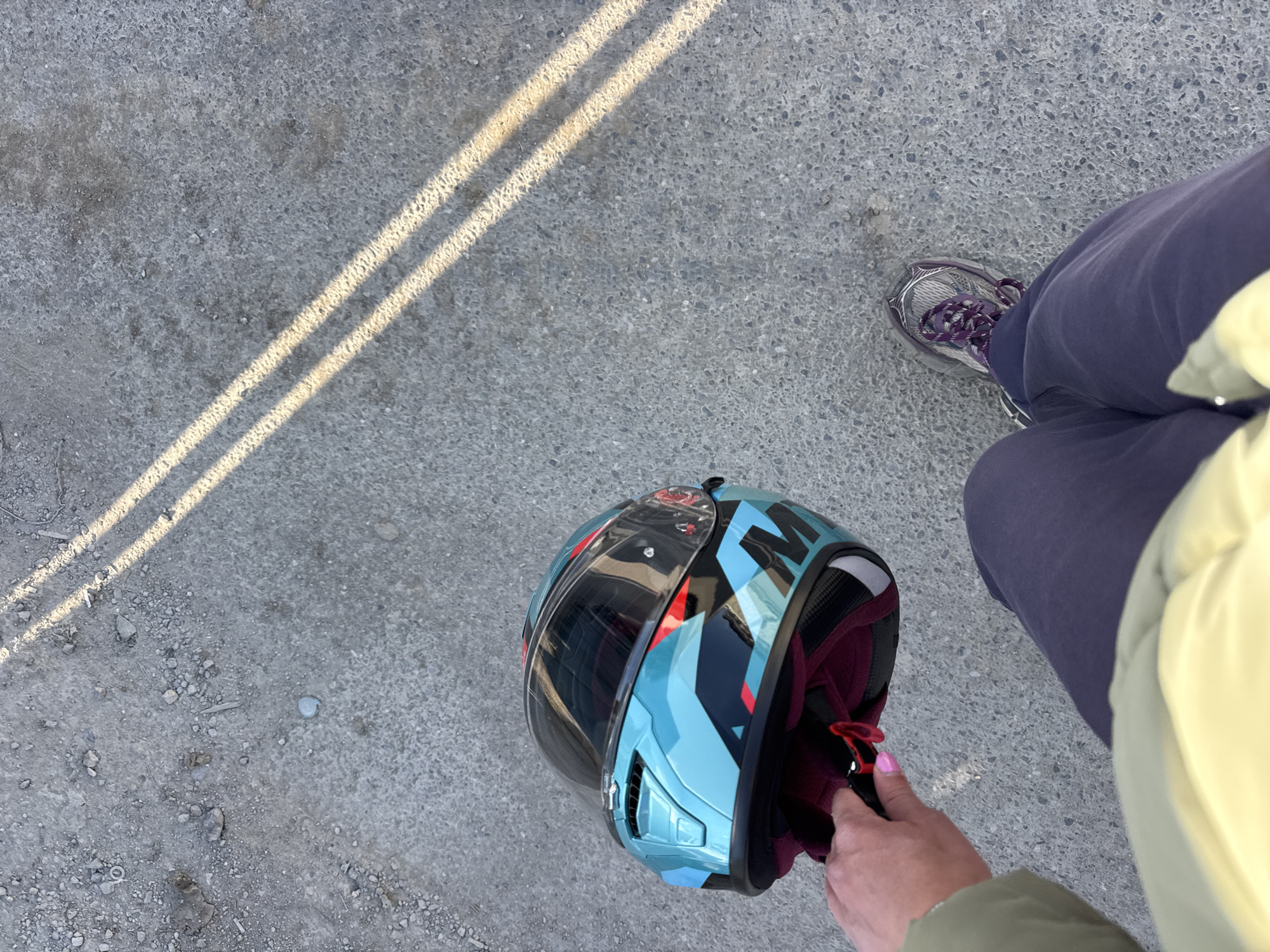 A high-angle shot shows a person standing on an outdoor concrete surface next to a motorcycle helmet. The person is wearing a dark grey running shoe, dark blue pants, and a light yellow jacket. The helmet is predominantly light blue with black, red, and white accents, and has a dark visor. The person's hand, with pink nail polish, is partially visible near the helmet's opening. Two parallel lines of light cross the concrete surface. The location is in an urban area, likely Sofia, Bulgaria, based on visual elements of the concrete and clothing.