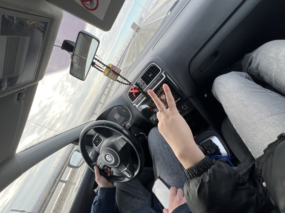 The image depicts the interior of a Volkswagen vehicle traveling on a multi-lane highway in the vicinity of Khapry, Russia, under an overcast sky. The driver's left hand, clad in a dark blue sleeve, grips the black steering wheel. Seated in the front passenger seat, an individual's right hand, wearing a ring on the fourth finger, is extended towards the camera in a "peace sign" gesture. This passenger is wearing a black jacket and light grey jeans. Their left hand holds a white smartphone on their lap. The black dashboard features an infotainment screen displaying Cyrillic text, and air vents with a red 'X' charm attached. A beaded air freshener, marked with "STARBOX" and Cyrillic text, hangs from the rearview mirror. Outside the vehicle, the highway shoulders exhibit patches of snow or ice, and guardrails line the road. A white truck is visible in the distant right lane, and the horizon reveals what appear to be industrial structures under a bright, cloudy sky.