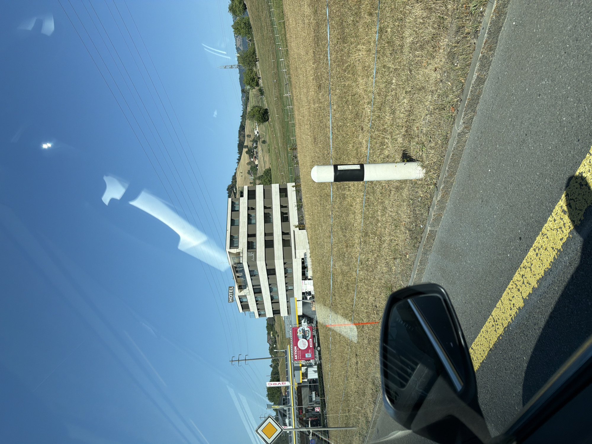 This is a horizontal, eye-level, outdoor photograph taken from a vehicle, showing a modern hotel building and a gas station in Immensee, Switzerland. The sky is a clear, bright blue with some sun glare and lens flare artifacts visible, particularly in the upper portion. A few power lines stretch across the sky.
In the mid-ground, a multi-story hotel with a beige facade and dark windows is prominently featured. The word "HOTEL" is visible in large letters on the roof. To the left of the hotel is a gas station with white and red awnings. A white truck with a red advertisement banner showing a meal is parked at the gas station. A diamond-shaped yellow and white road sign stands near the gas station entrance.
The foreground shows a section of a road with a broken yellow line painted on it. A car's side mirror is visible in the lower left corner, reflecting a blurred image of the hotel. Beyond the road is a dry, grassy field with a thin wire fence and a white post with black markings. In the background, rolling green hills with scattered trees and houses are visible, characteristic of the Swiss landscape. The overall scene depicts a bright, sunny day in a semi-rural or suburban area.