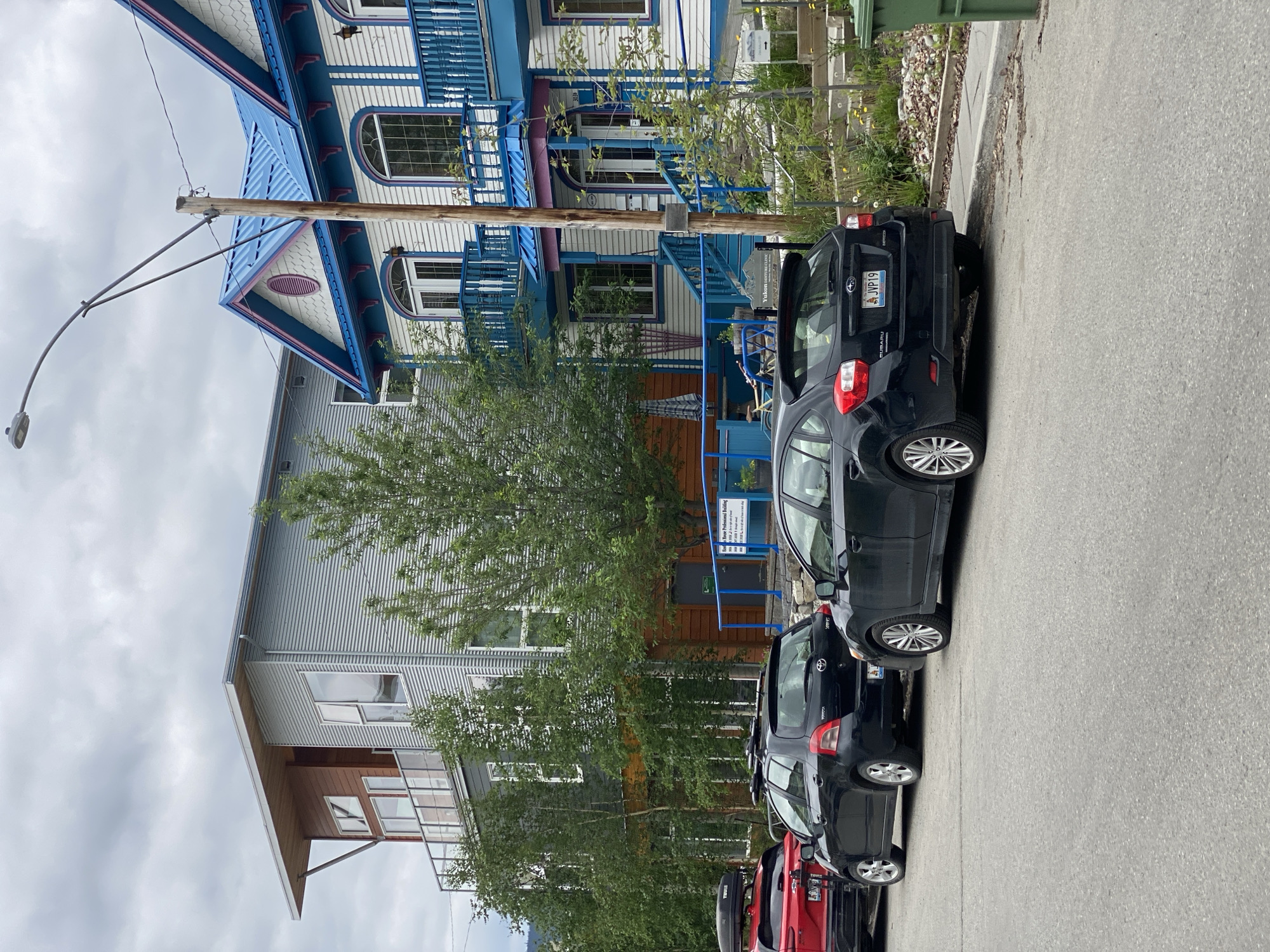 The image depicts a street scene in Whitehorse, Canada, showcasing a row of buildings and parked vehicles. On the right side of the frame, a historic-looking building with white siding, blue trim, and a bright blue roof stands prominently. Decorative blue railings adorn its balconies and staircases. To the left of this building is a more modern structure with corrugated metal siding and a flat roof, featuring large windows and a balcony.
Several cars are parked along the street. In the foreground, a black Subaru hatchback is parked facing away from the camera. To its left, a black Toyota hatchback is also parked. Further down the street, a red SUV is visible. The street itself is paved asphalt, and a sidewalk runs along the right side of the frame, bordered by a grassy area with some vegetation. A green garbage bin is situated on the sidewalk. A street lamp with a curved arm is visible overhead, attached to a wooden utility pole. The sky is overcast with cloudy conditions. There are no people visible in the image. The scene appears to be a commercial or residential area within the city.