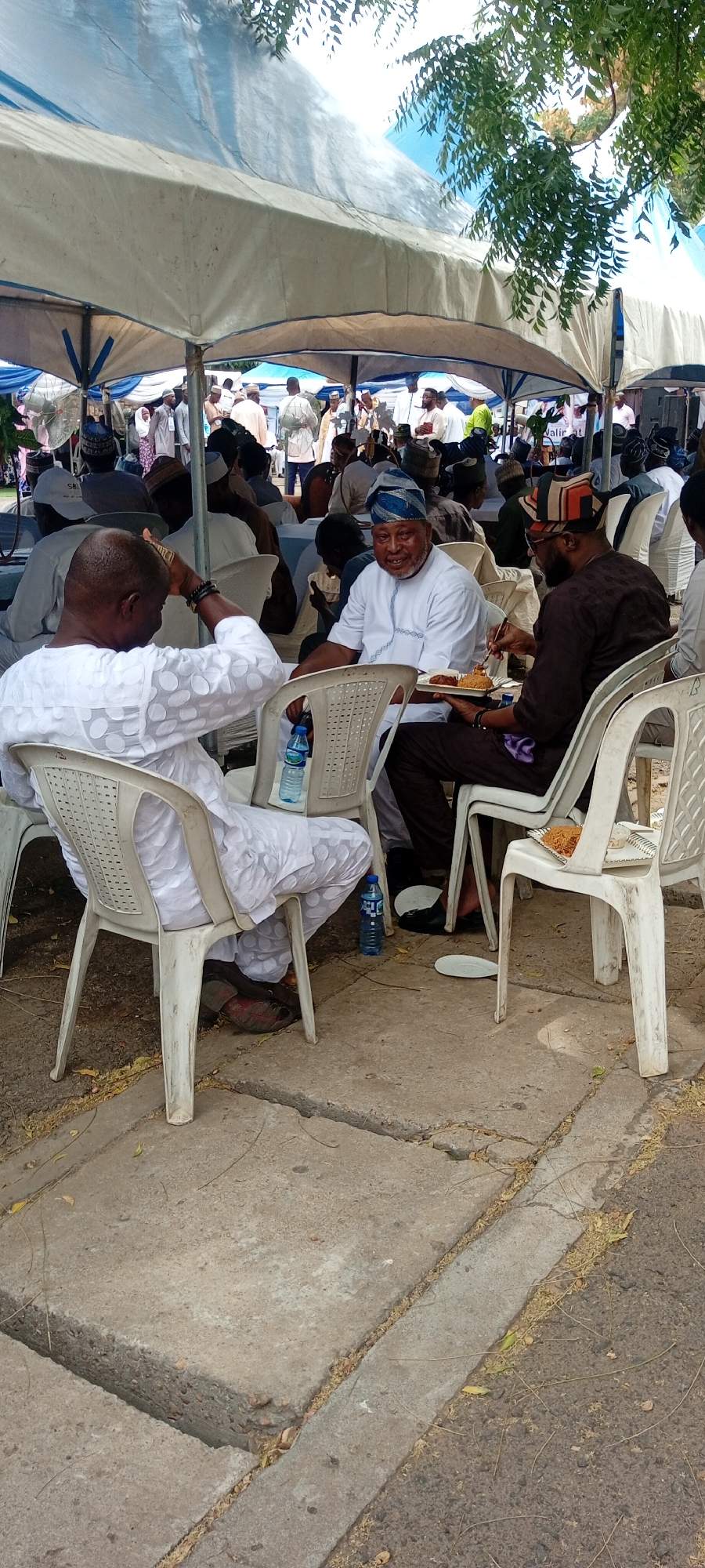 The scene captures a gathering of people in Goron Dutse, Nigeria. The individuals are seated on white plastic chairs under a large white tent, possibly for an event or celebration. Three men are prominently featured in the foreground: one dressed in white with a beaded bracelet, another in a white shirt and a blue patterned cap, and a third in a brown attire with a cap. They are all eating food from plates. There are water bottles near them. In the background, more people are seen milling around under the tent. The setting appears to be outdoors, with a concrete surface and some foliage visible near the edge of the tent.