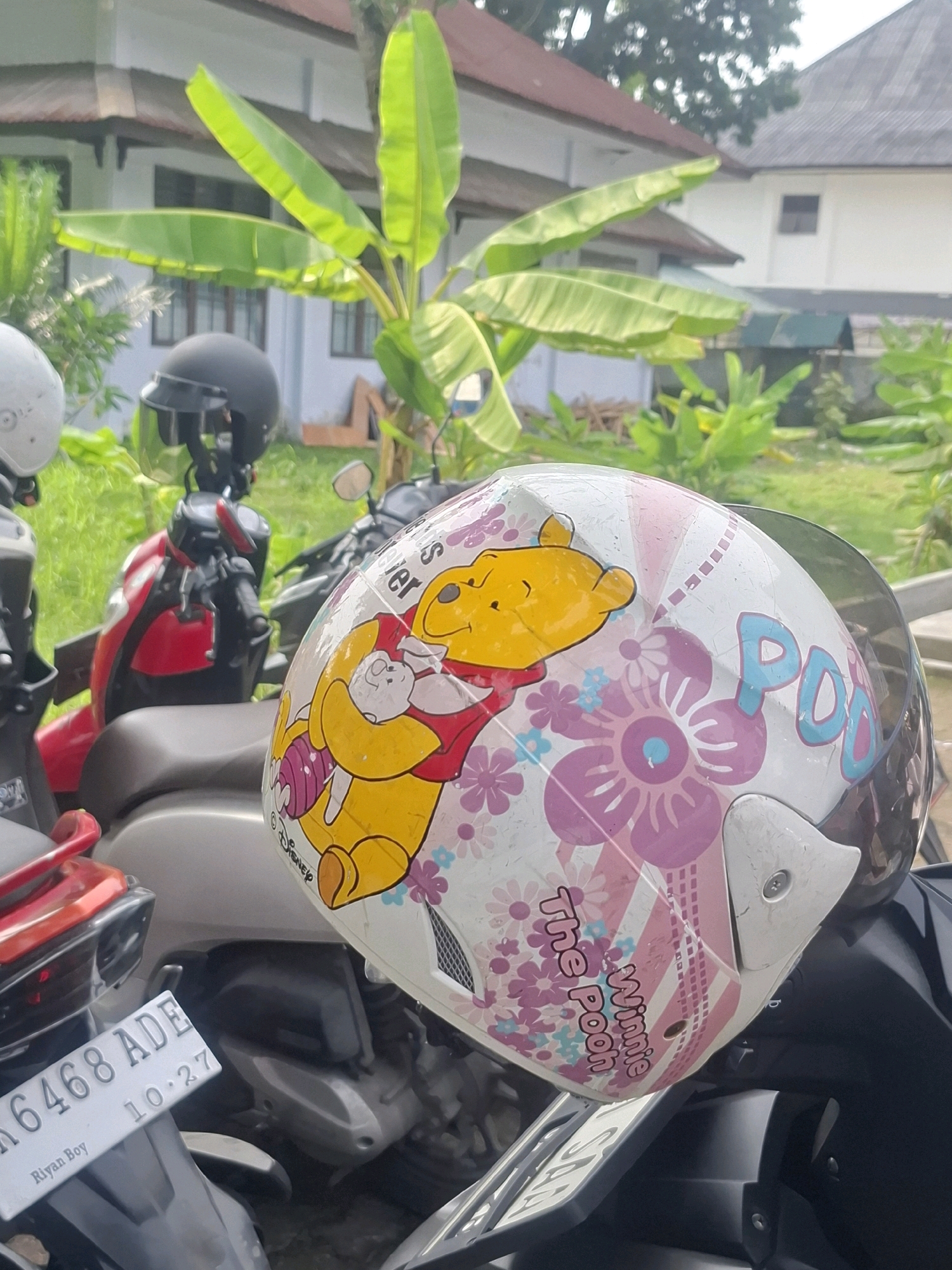 In Medan Estate, Indonesia, an outdoor scene unfolds with multiple motorcycles parked under the shade of lush green foliage, likely banana trees, with a building visible in the background. The focal point is a white helmet adorned with a vibrant cartoon design of Winnie the Pooh and Piglet, surrounded by pink and blue flowers and stripes. The helmet's visor is partially down. To the left of this helmet, another black helmet is visible on a red and black motorcycle. A license plate reading "6468 ADE" and "Riyan Boy" is partially visible on another motorcycle in the foreground. The ground appears to be a mix of dirt and grass. The overall atmosphere suggests a casual, everyday setting in a tropical environment.