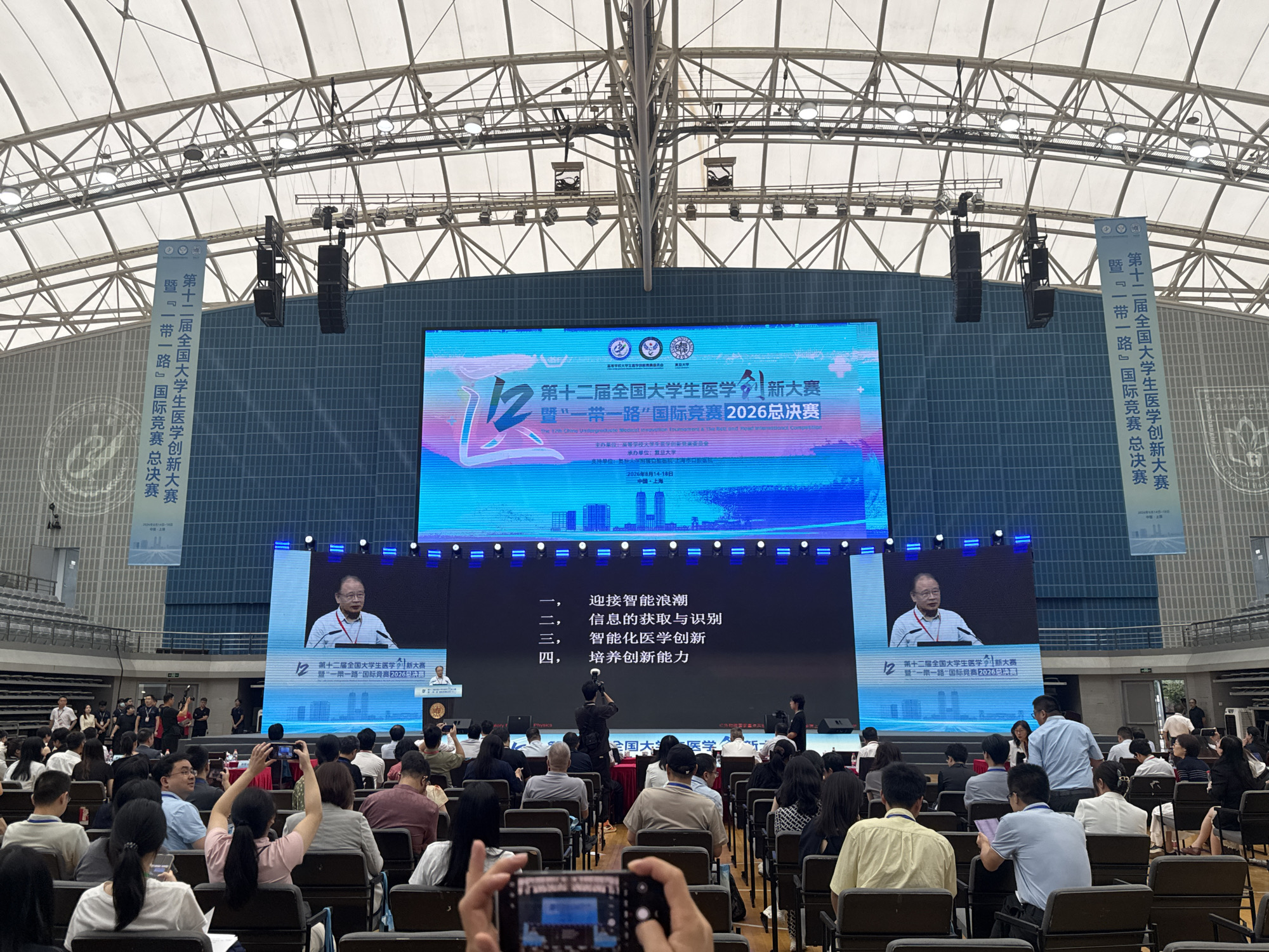 This image depicts an indoor event, likely a conference or competition, held in Yangpu District, China. A large LED screen dominates the stage, displaying text and images related to an event titled "The 12th National University Student Medical Innovation Competition" and "Belt and Road" International Competition, with the year 2026. Two men are visible on the screen, one speaking at a podium. On stage, a woman stands to the right of the screen. In the audience, a crowd of people are seated, many holding up their phones to take pictures or videos of the event. The environment appears to be a large hall or gymnasium with tiered seating. Flags bearing Chinese text are displayed on either side of the main screen. The ceiling is a complex structure of metal beams and white fabric, with lighting fixtures visible.