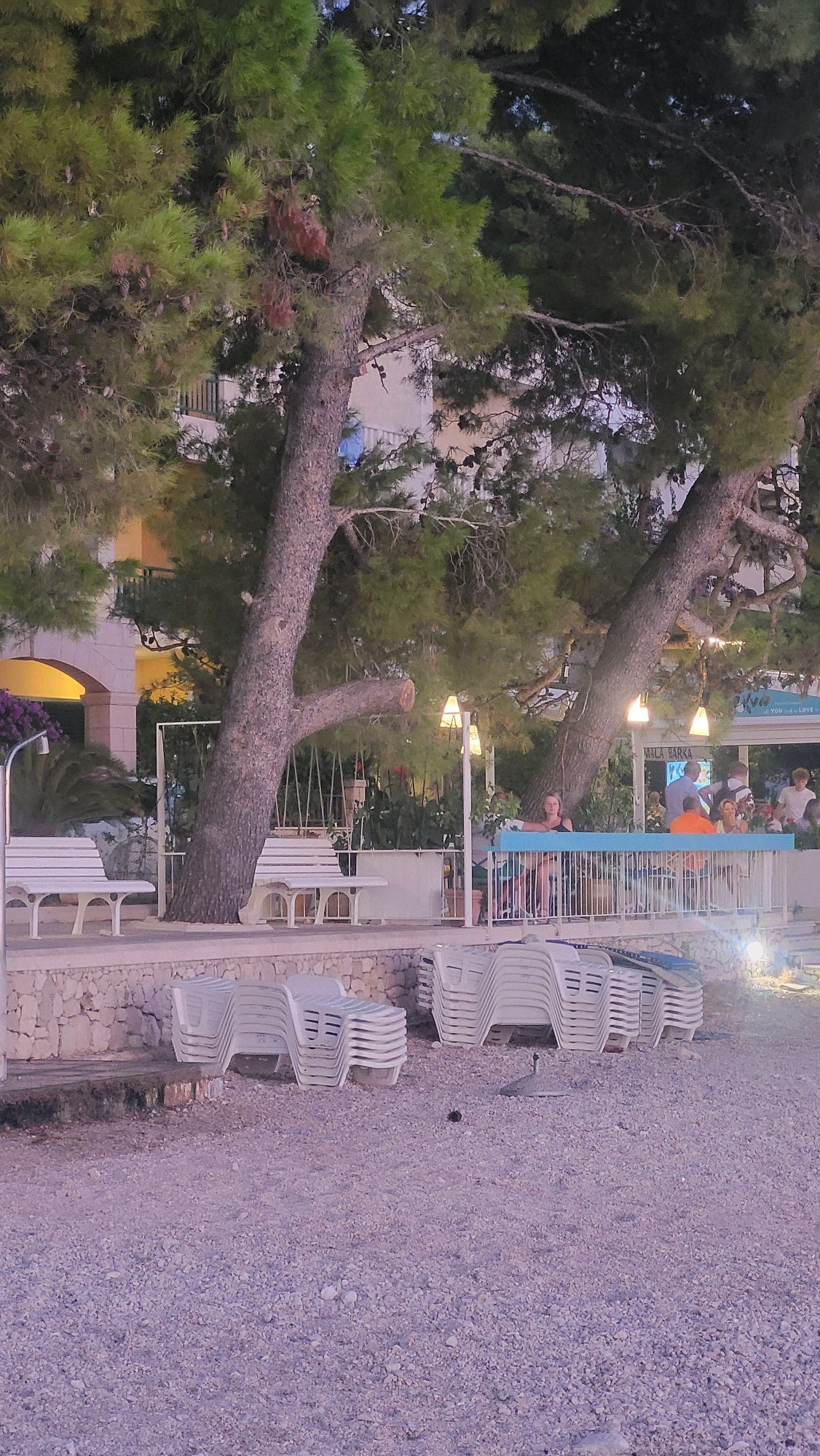 The image depicts a view of a beach area in Općina Tučepi, Croatia, likely in the evening given the lighting. In the foreground, stacks of white plastic lounge chairs are visible on a pebble beach. Behind the chairs, a stone wall separates the beach from a paved area with more white benches. Large trees with dense green foliage dominate the midground, with their trunks and branches casting shadows. Several white outdoor lamps are suspended from the trees. Further back, a building with a light-colored facade and arched windows is visible. A covered outdoor seating area with a blue railing is also present, where several people are seated and standing. Some individuals are conversing, while others appear to be dining. A sign above the seating area reads "Mala Barka". The overall scene suggests a relaxed atmosphere at a coastal establishment.