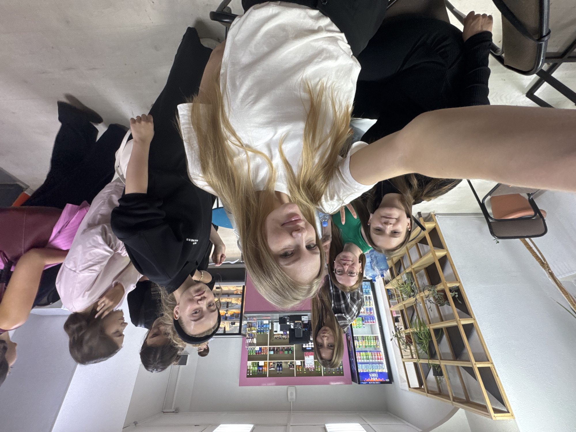 A group of six young women are posed for a selfie in what appears to be a cafe or small store in Perm, Russia. The photo is taken from a high angle, slightly tilted. The interior has a pink color scheme. Shelves stocked with drinks and snacks can be seen in the background. A wooden structure is on the wall. The women are wearing casual clothing.