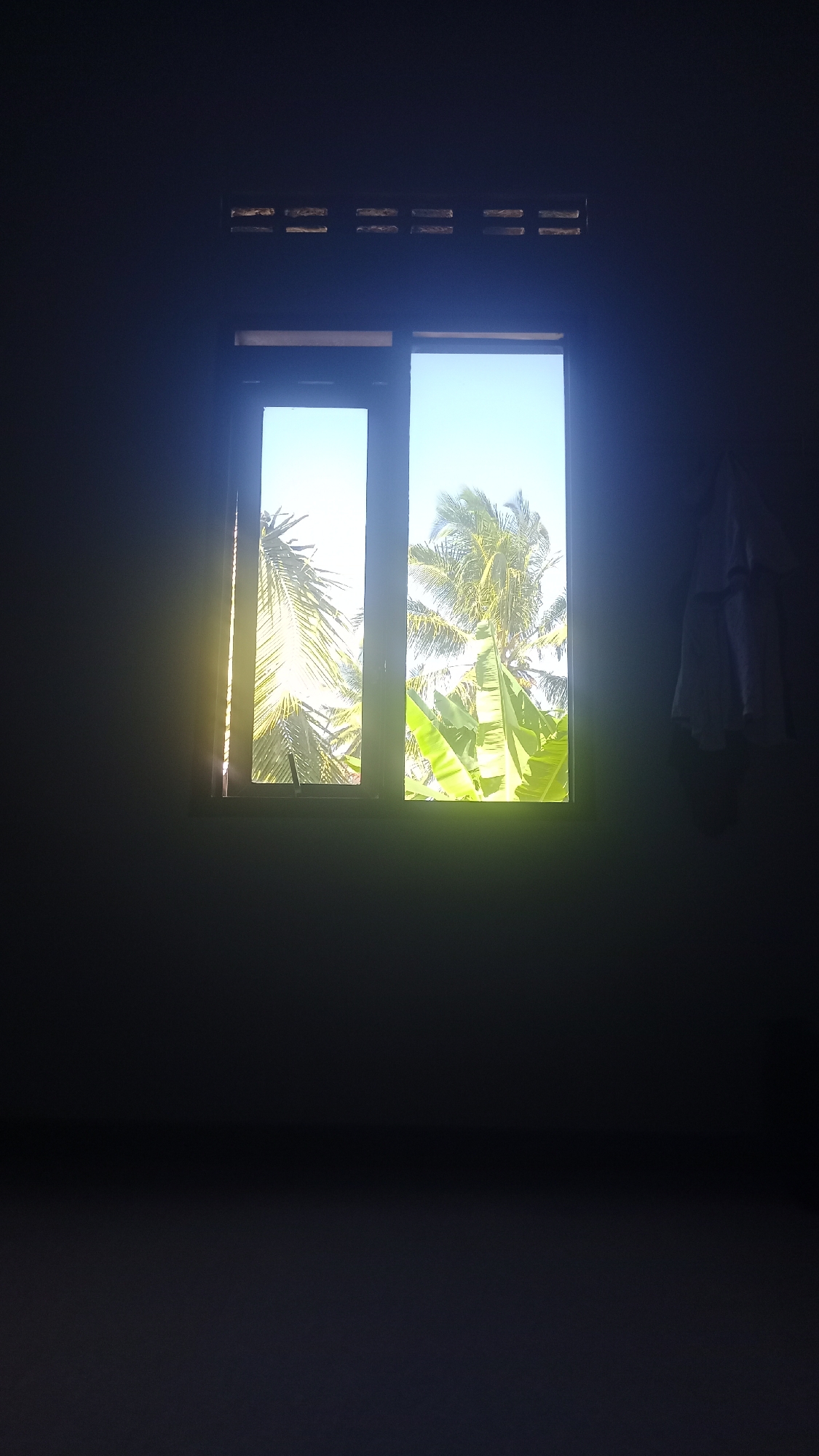 A vertical, dimly lit shot shows a double-paned window with a tropical view. The left pane is bathed in bright sunlight, creating a glare that obscures details. The right pane reveals a clear blue sky, palm trees, and large green banana leaves. Above the window, a row of small, rectangular vents spans the width of the frame. To the right of the window, a white garment hangs on the wall. The foreground and background of the room are cast in deep shadow, with the only light source being the window. This scene is located in Pakemitan, Indonesia.