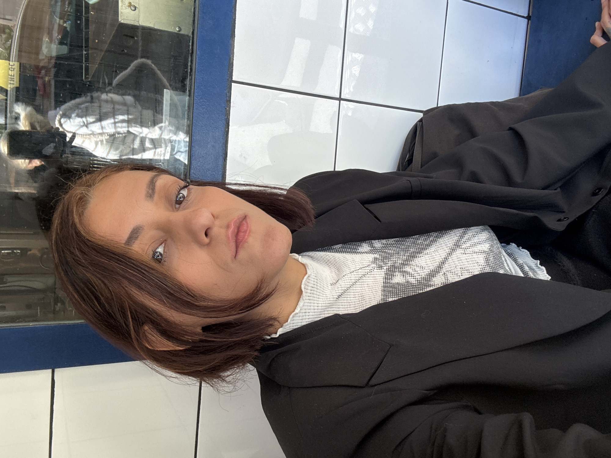 A medium shot depicts a woman with light brown skin and light brown hair with reddish-brown streaks, wearing a black blazer over a white patterned top. She is positioned indoors, potentially in a subway station, as evidenced by the white tiled walls with thin black grout lines and a blue border near a window or display case. The reflections in the window suggest the presence of other people and objects outside. The overall impression is of an urban setting, possibly Barcelona, Spain, inferred from the subway station backdrop, a common form of public transportation.