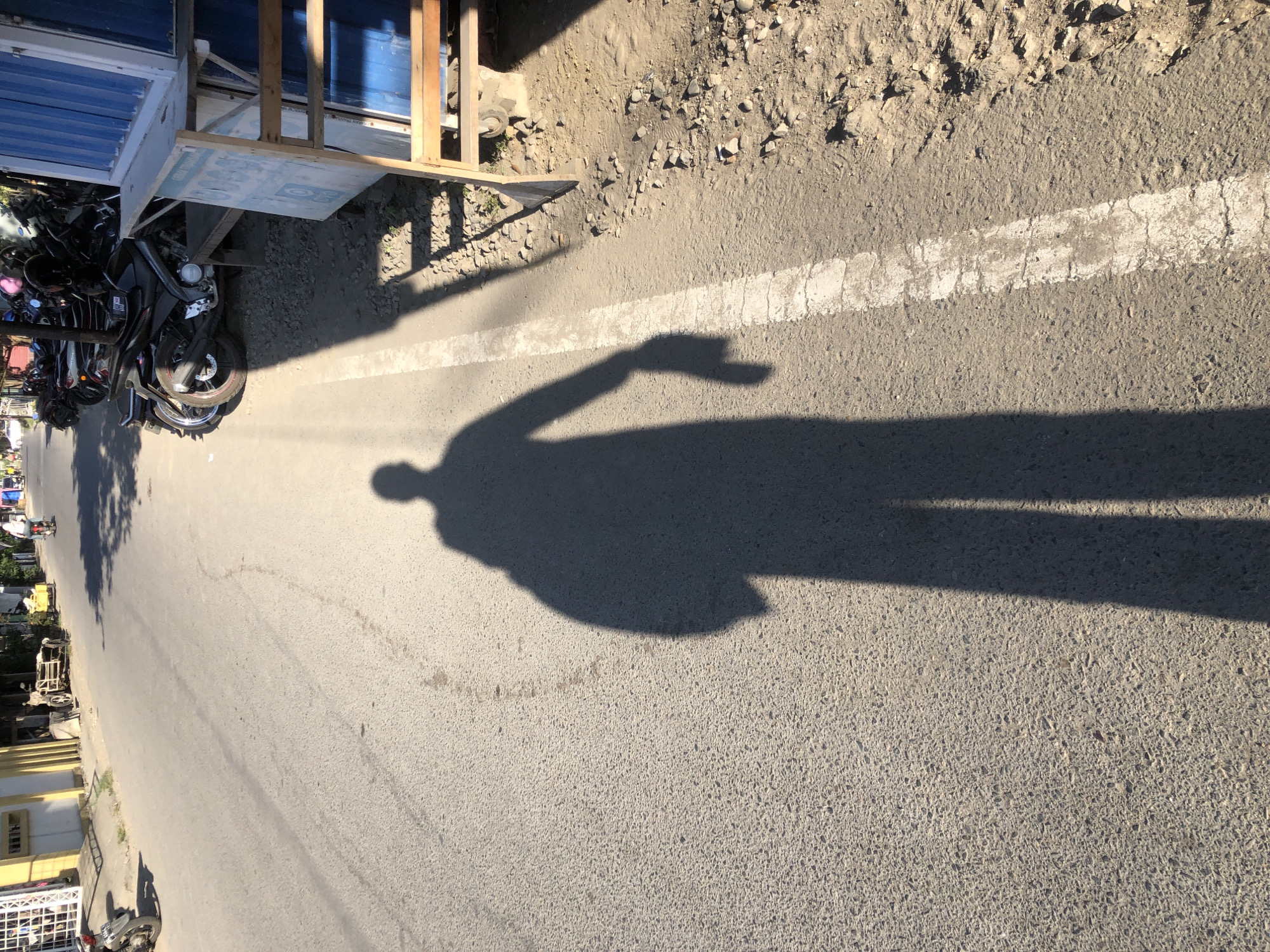 The image shows a sunlit asphalt road in Lhokseumawe, Indonesia. The road has a white line marking. The shadow of a person is cast on the road, with one arm raised. A row of parked motorbikes is visible on the side of the road, adjacent to a structure with a blue awning and wooden frame. Vegetation and buildings are seen on the opposite side of the road. The surface beside the road is covered with gravel.