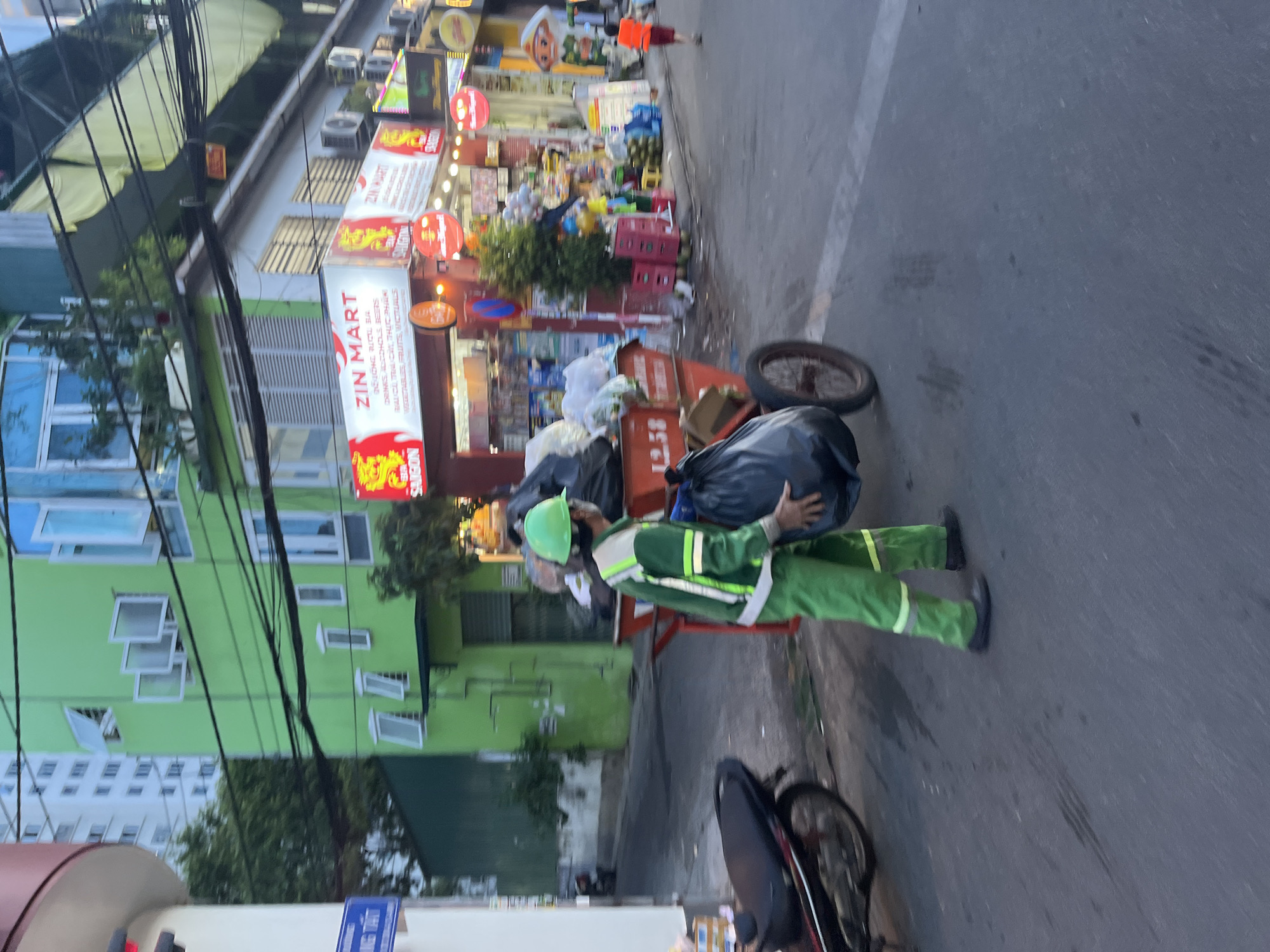 A street scene in Bac Nha Trang Ward, Vietnam, depicts a sanitation worker in a green uniform and helmet lifting a large black trash bag onto a wheeled cart. The background shows a commercial area with storefronts, including one labeled "ZIN MART," identifiable by its signage and visible merchandise. The street is paved, and the sky is overcast, suggesting either early morning or late afternoon lighting conditions. A motorbike is partially visible in the foreground.