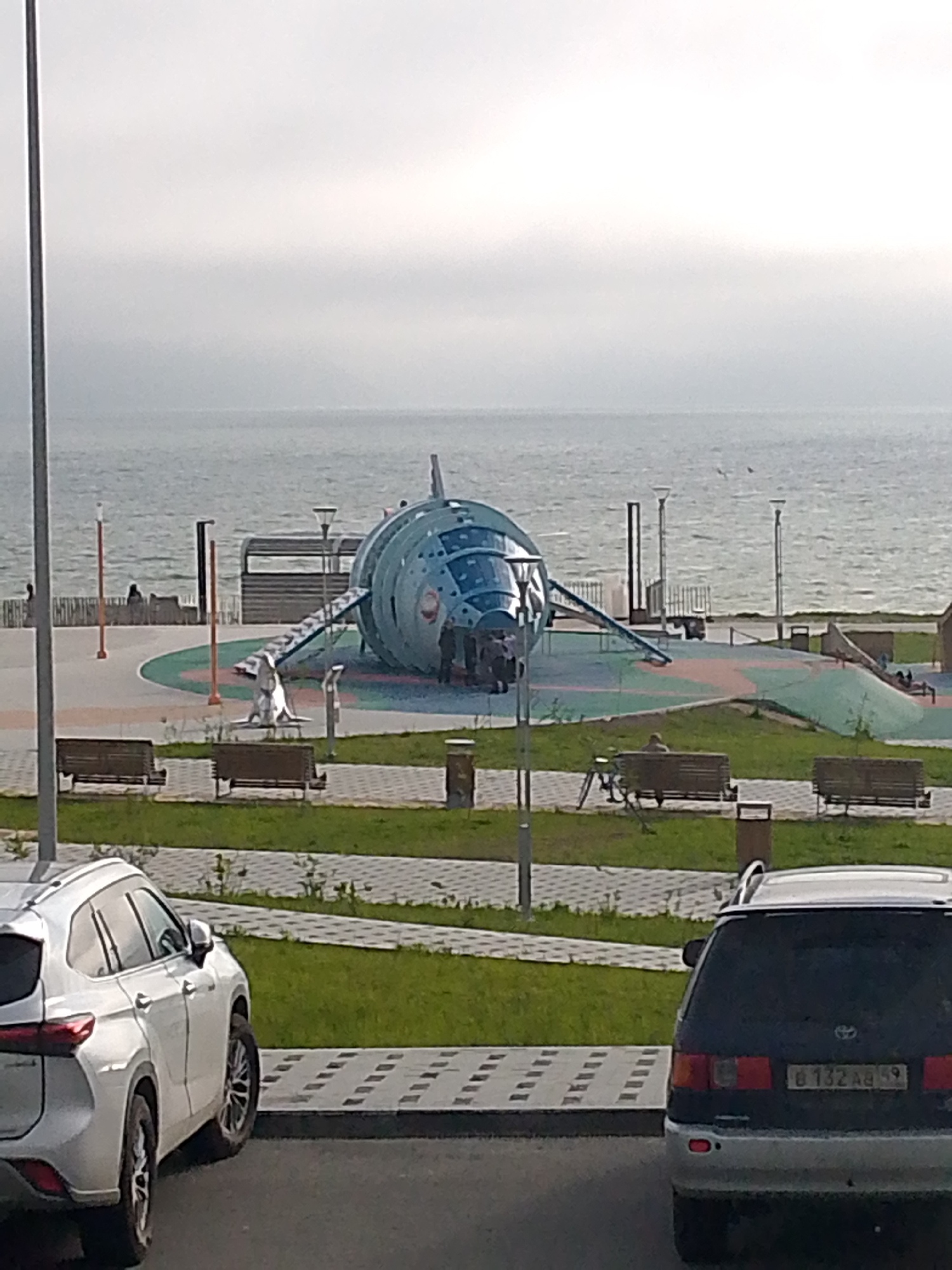 The image shows a playground designed to look like a rocket or spaceship, situated near a body of water, likely a sea or large lake, under an overcast sky. The playground structure is large and metallic blue, with windows and a ramp leading up to it. Several people are visible near the structure, some entering or exiting. In the foreground, two cars are parked: a white SUV on the left and a dark minivan on the right. The minivan has a license plate that appears to read "B 132 AA 59". The area around the playground is paved with patterned tiles and grass, with benches and trash receptacles present. In the background, a railing separates the paved area from the water, with a few distant figures visible on the other side of the railing. The overall setting suggests an outdoor recreational area in a coastal or lakeside city. The license plate suggests the location is in Russia. Given the context of a prominent spaceship-themed playground near the sea in Russia, this scene is likely located at the "Navigator" Space-themed Playground in Magadan, Russia, a well-known local attraction.