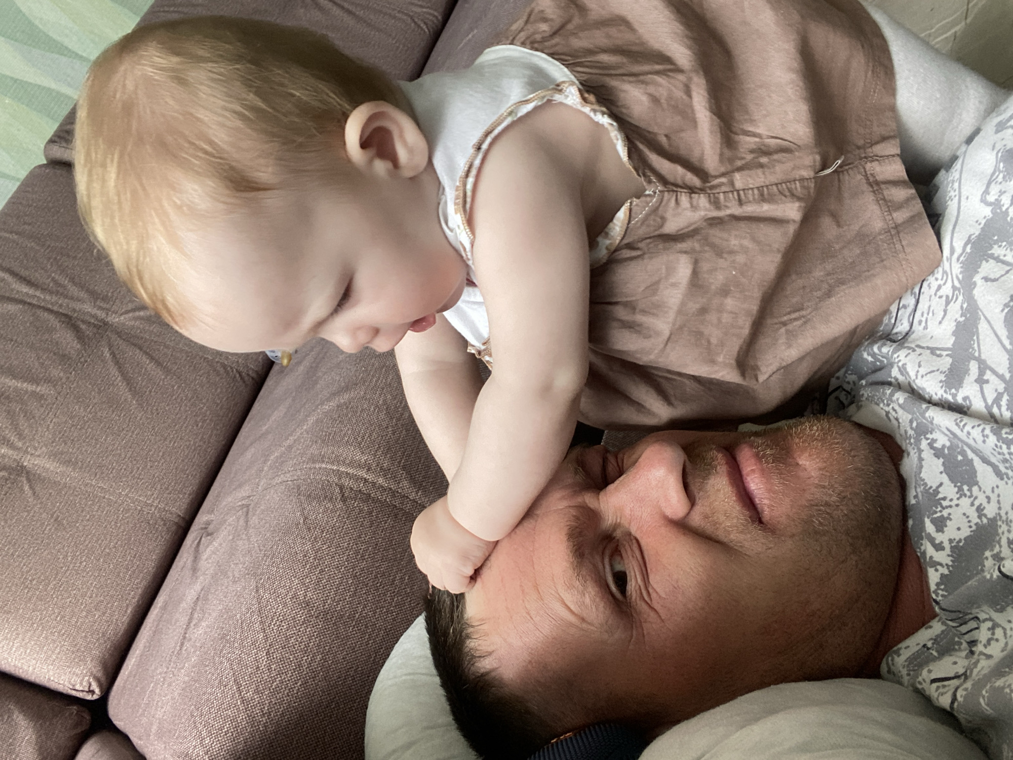 A close-up shot shows a light-skinned man lying on a maroon-colored upholstered couch, while a baby, wearing a brown and white dress, interacts with his face. The baby is positioned above him and appears to be touching his forehead with their hand. The man's eyes are open, and he seems to be looking at the baby. The background includes a white pillow and a patterned grey blanket. No specific information confirms the location as Фоки, Russia.