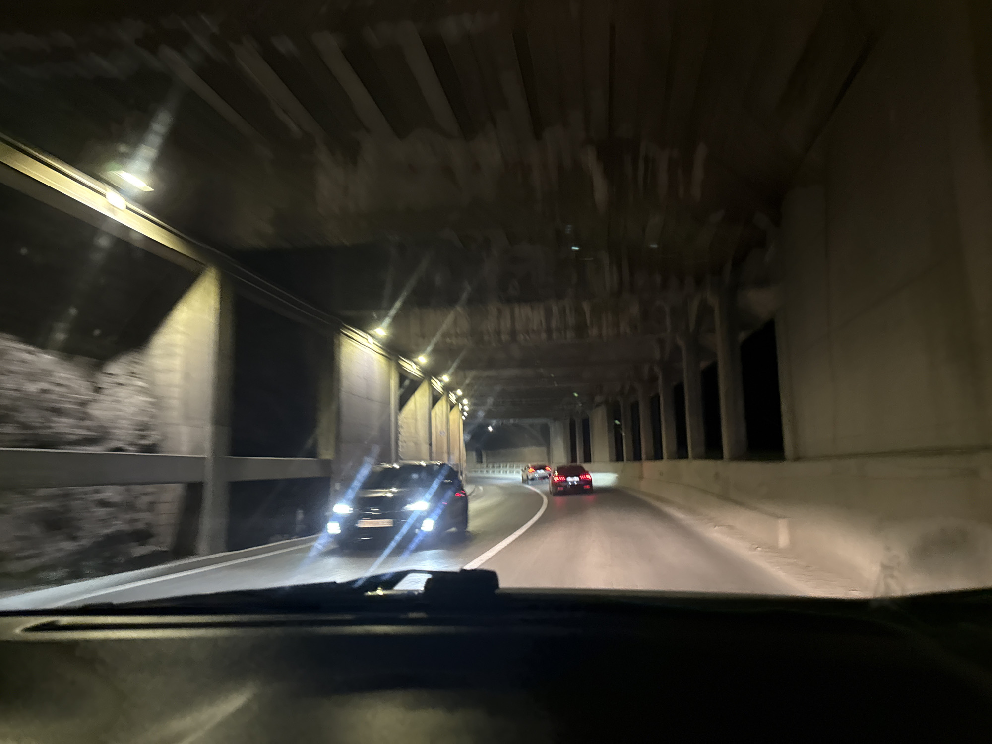 The image shows a view from inside a car, driving through a tunnel. Other cars are visible ahead, including one with bright headlights. The tunnel structure is made of concrete with visible beams and columns. The location is likely near the city of Budva, Montenegro, given the visual similarity to images of tunnels in that area.