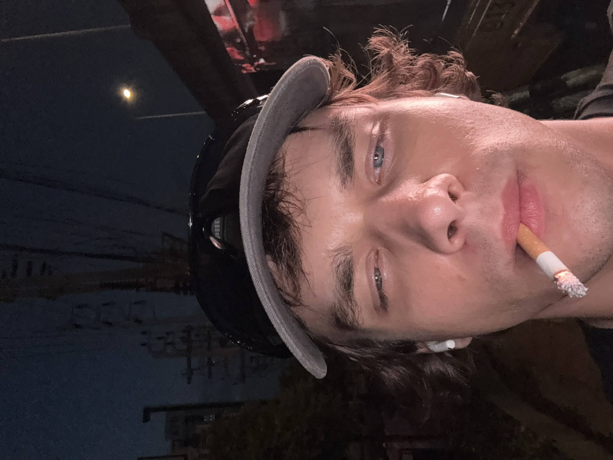 A close-up, Dutch-angled portrait captures a young man outdoors at night in Bangkok, Thailand. The man, likely in his 20s or 30s, has light skin, bright blue eyes, and curly brown hair. He wears a black helmet, partially covered by a grey, ribbed fabric brim that is pushed back on his head.
He is actively smoking, with a lit cigarette held between his lips, showing a visible column of ash. A white earbud is noticeable in his left ear. His skin has a slight sheen, suggesting warmth or recent activity.
The setting is distinctly nocturnal, with a dark sky dominating the upper left portion of the image, where a small, bright crescent moon is visible. The background is blurred, hinting at an urban environment with indistinct structures and overhead wires, typical of a city. No discernible text is visible in the image.