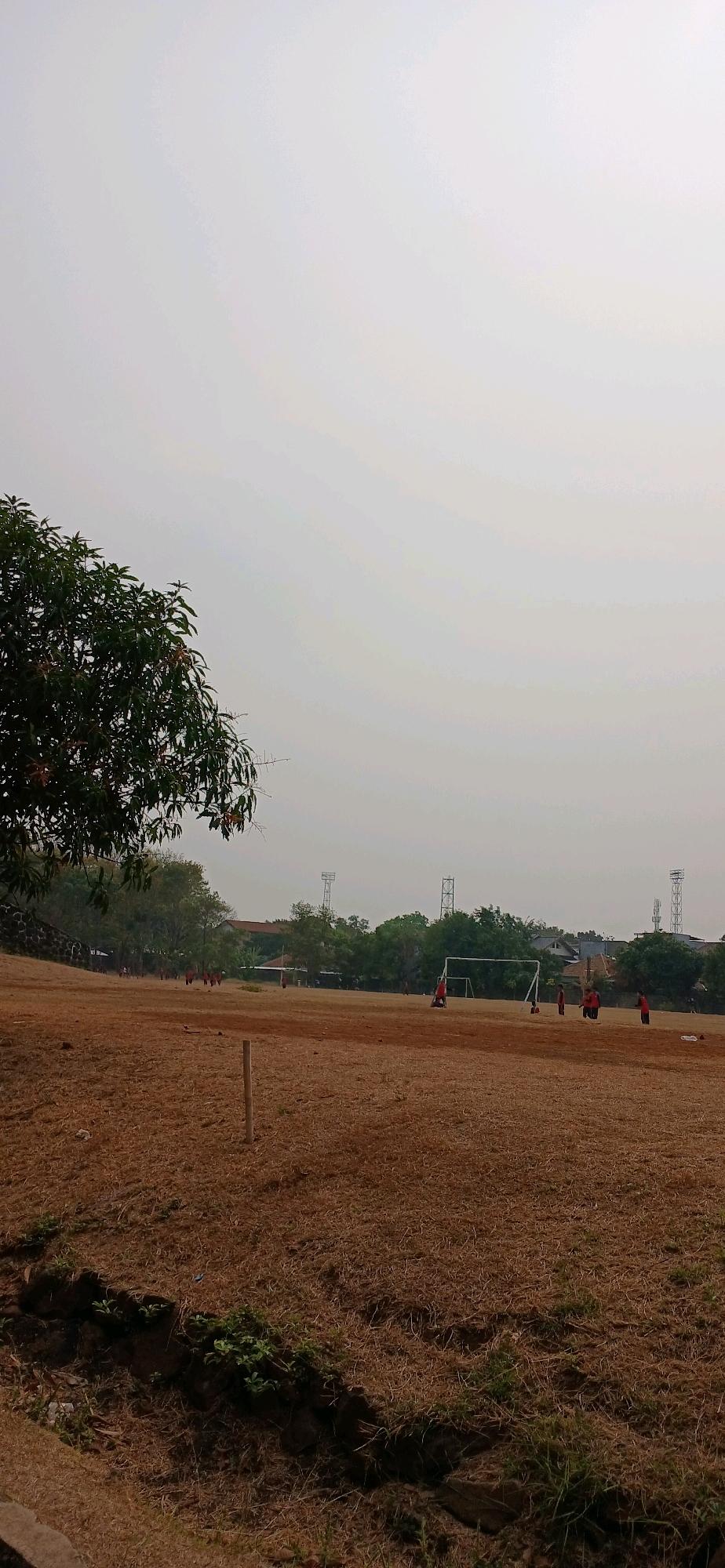 A wide shot of a dry, brown soccer field under a hazy, overcast sky in Cirebon, Indonesia. Several figures, likely young people wearing red shirts, are spread across the field, some near a soccer goal. In the foreground, the grass is patchy and dry. A single wooden post stands in the middle of the field. In the background, trees, buildings, and two tall transmission towers are visible. The overall impression is of a sparsely populated, somewhat neglected public space.