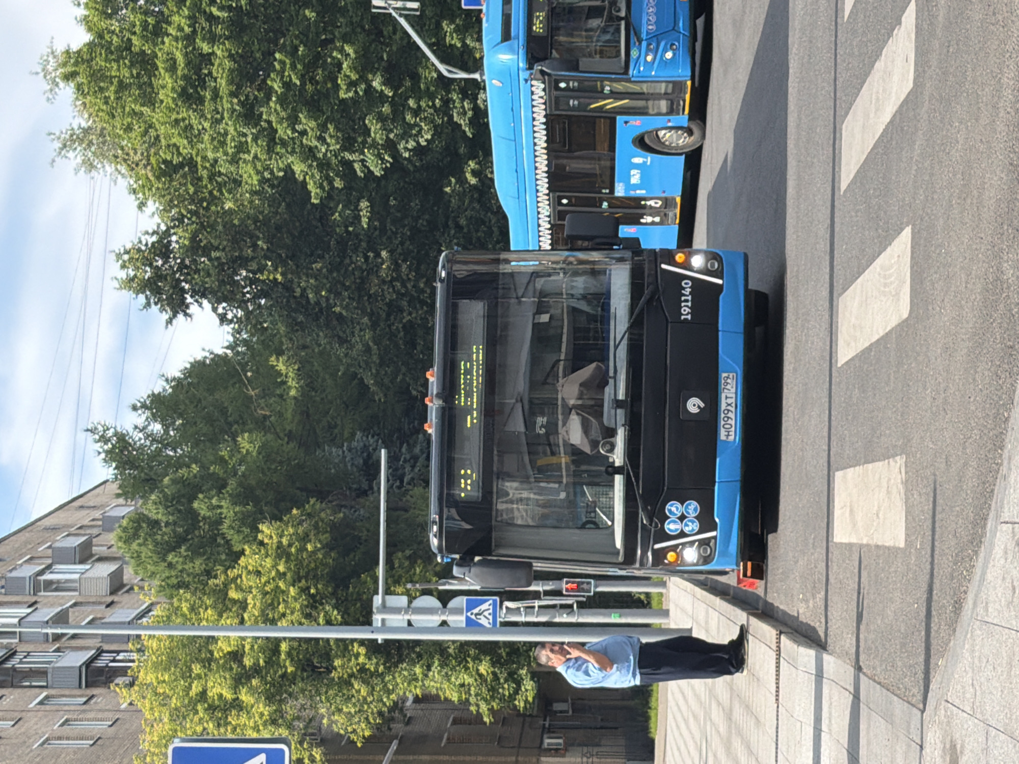 A man in a blue shirt and dark pants stands on a sidewalk, facing away from the camera and looking at his phone. He is positioned next to a light pole with a pedestrian crossing sign. A black and blue bus is parked on the street in front of him, partially obscuring the view of another blue bus behind it. The bus in the foreground has its headlights on and displays a license plate that reads "H099XT 799". The environment appears to be an urban street with trees lining the background and a building on the left side of the image. The sky is partly cloudy.
