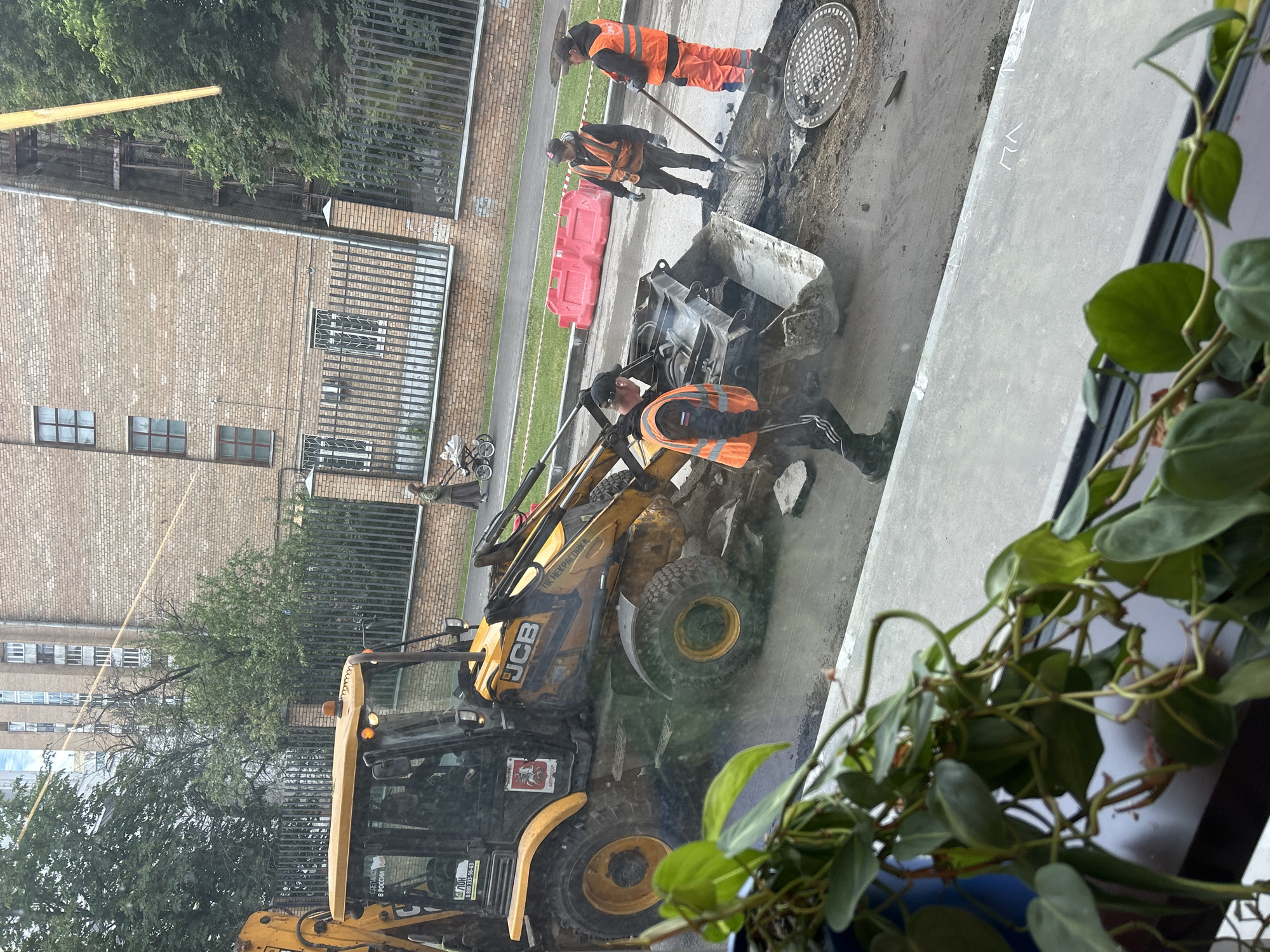 The image depicts a street repair scene in an urban environment, possibly Unknown, Unknown. A yellow JCB backhoe loader is positioned in the foreground, with a construction worker in an orange safety vest appearing to interact with the machinery. Two additional workers, also in orange vests, are near a manhole, one holding a shovel. Debris and excavated material surround the work area. A brick building is in the background with a metal fence. A potted plant obscures part of the bottom right corner of the view, suggesting the image was taken from indoors.