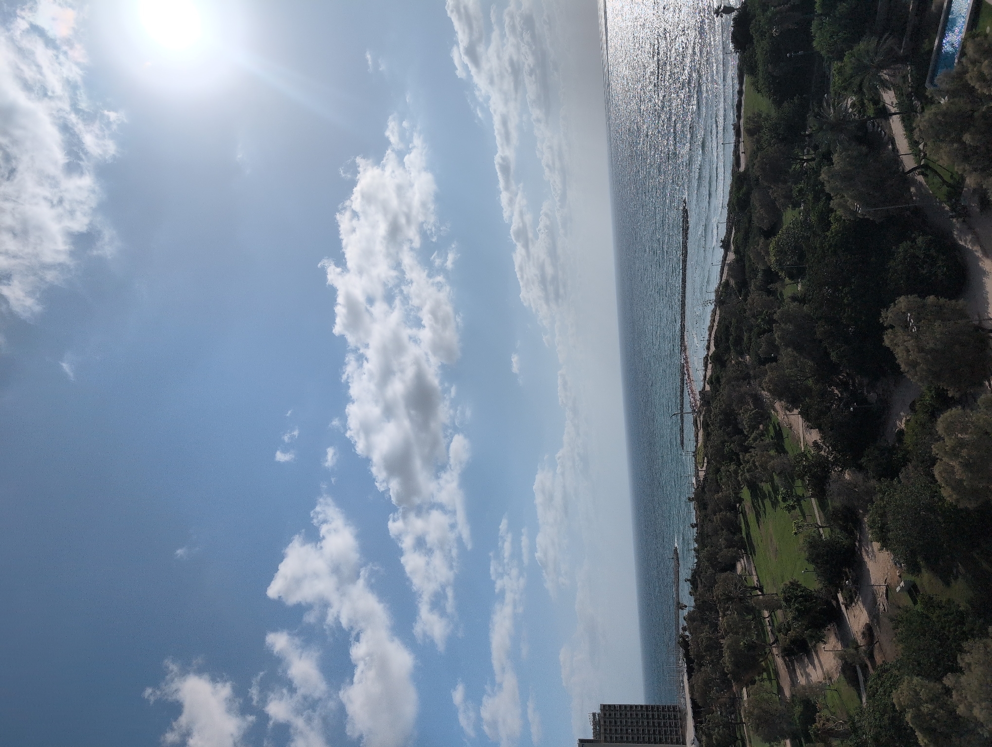 This image is an elevated view of a park and coastline in Tel Aviv, Israel, likely taken from a tall building. The sky is a clear blue with scattered white clouds, and the sun is shining brightly in the upper right corner. The sea is a deep blue, with the sunlight reflecting off its surface, creating a shimmering effect.
In the foreground, a park area is visible, with green lawns, trees, and pathways. There are scattered trees with dense foliage, as well as some with sparser leaves, suggesting a mix of species and possibly varying seasons. There appear to be some park benches visible in the shaded areas. A bright blue swimming pool with sunlight glinting off its surface is visible in the lower right corner of the park.
The middle ground shows the coastline, with a promenade or walkway running along the edge of the park. There are people visible on the beach and in the water, some appearing to be sunbathing or swimming. A pier or breakwater extends into the sea, with a lighthouse or structure at its end.
In the background, a tall, modern-looking building, likely an apartment or office building, is situated on the left side of the frame, overlooking the park and the sea. The overall scene depicts a pleasant day at the beach and park in a coastal city.