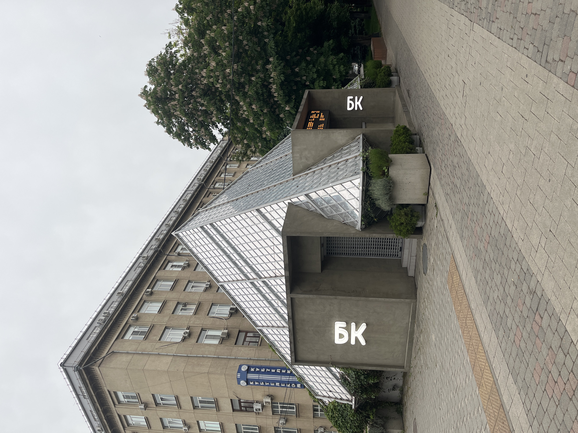 The image depicts a view of the 'Crystal Music Hall' in Krasnodar, Russia. A modern glass structure, resembling a pyramid, adjoins an older, multi-story building with numerous windows and air conditioning units affixed to its exterior. The entrance to the hall is marked with the Cyrillic letters "БК" (likely standing for "Big Concert Hall"). A digital display showing the time is visible above one entrance. The surrounding environment includes lush trees and a paved pedestrian walkway.