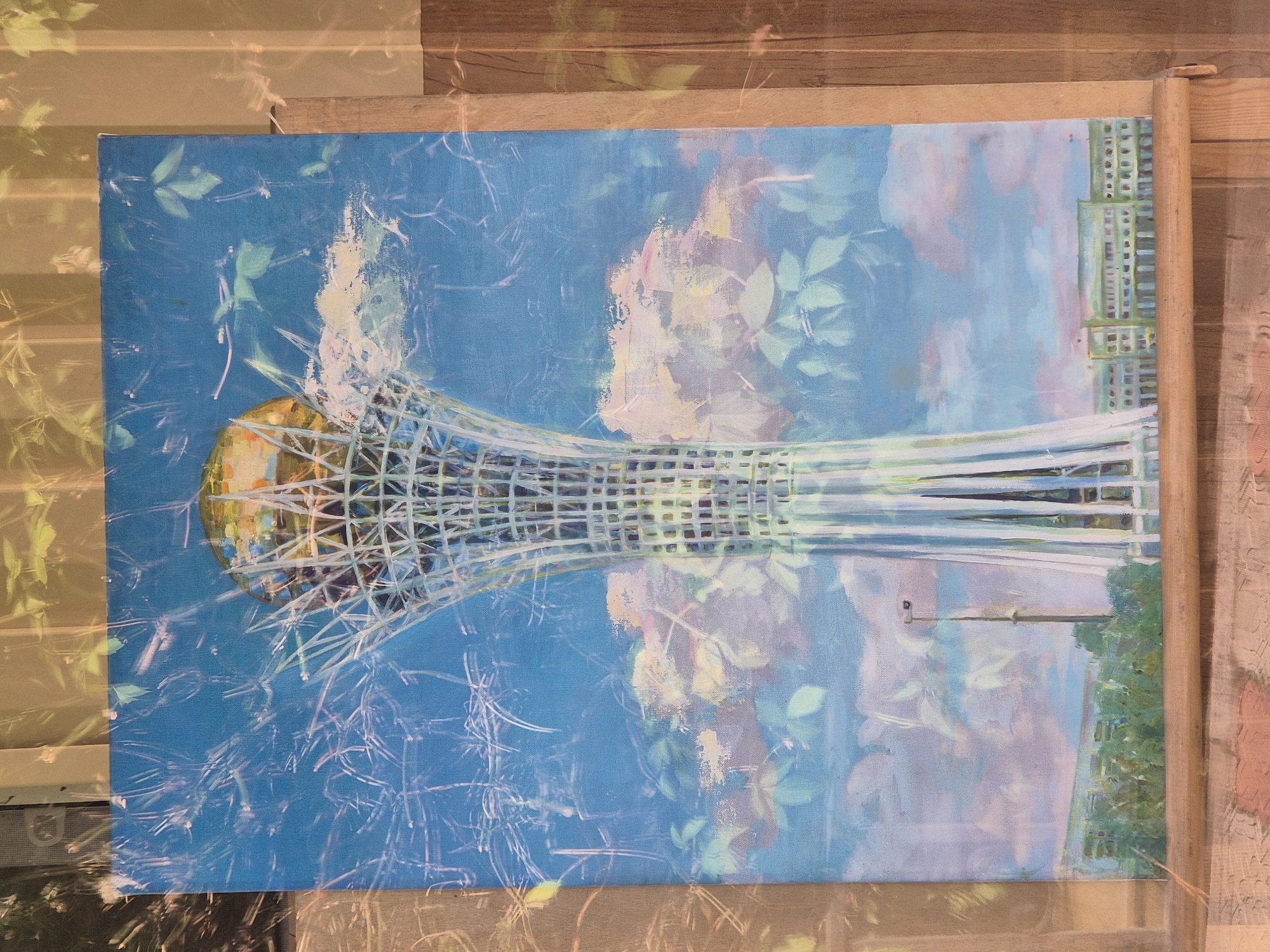 The image shows a painting of the Bayterek Tower in Astana, Kazakhstan. The tower is a prominent landmark in the city, and it is depicted in the painting with a golden globe at the top. The sky is blue with some clouds, and there are trees and buildings in the background. The painting is framed by wooden edges, and there are reflections of leaves and branches on the surface of the painting, suggesting it is displayed in a setting with natural light. The overall style of the painting is impressionistic, with visible brushstrokes and soft colors.