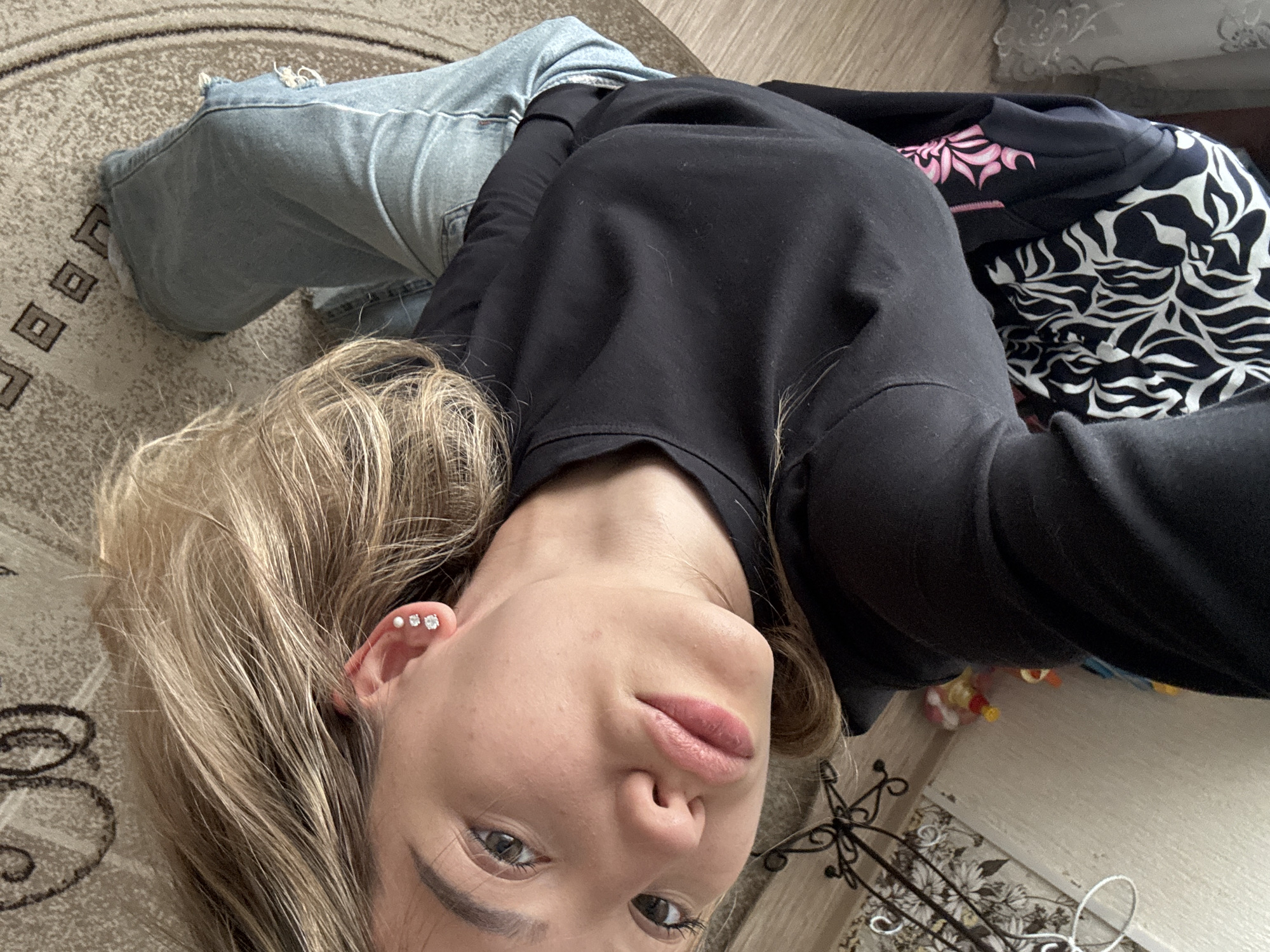 A woman with blonde hair is lying on a rug in an apartment in Noviy, Russia. She is wearing a black sweatshirt and blue jeans. She is looking up at the camera, and her eyes are open. She has two piercings in her left ear. The rug has a circular pattern with brown and beige colors. In the background, there is a chair with a black and white patterned fabric, a floor lamp, and a wall with wallpaper. There are also some toys on the floor. The image is a selfie taken from a high angle.