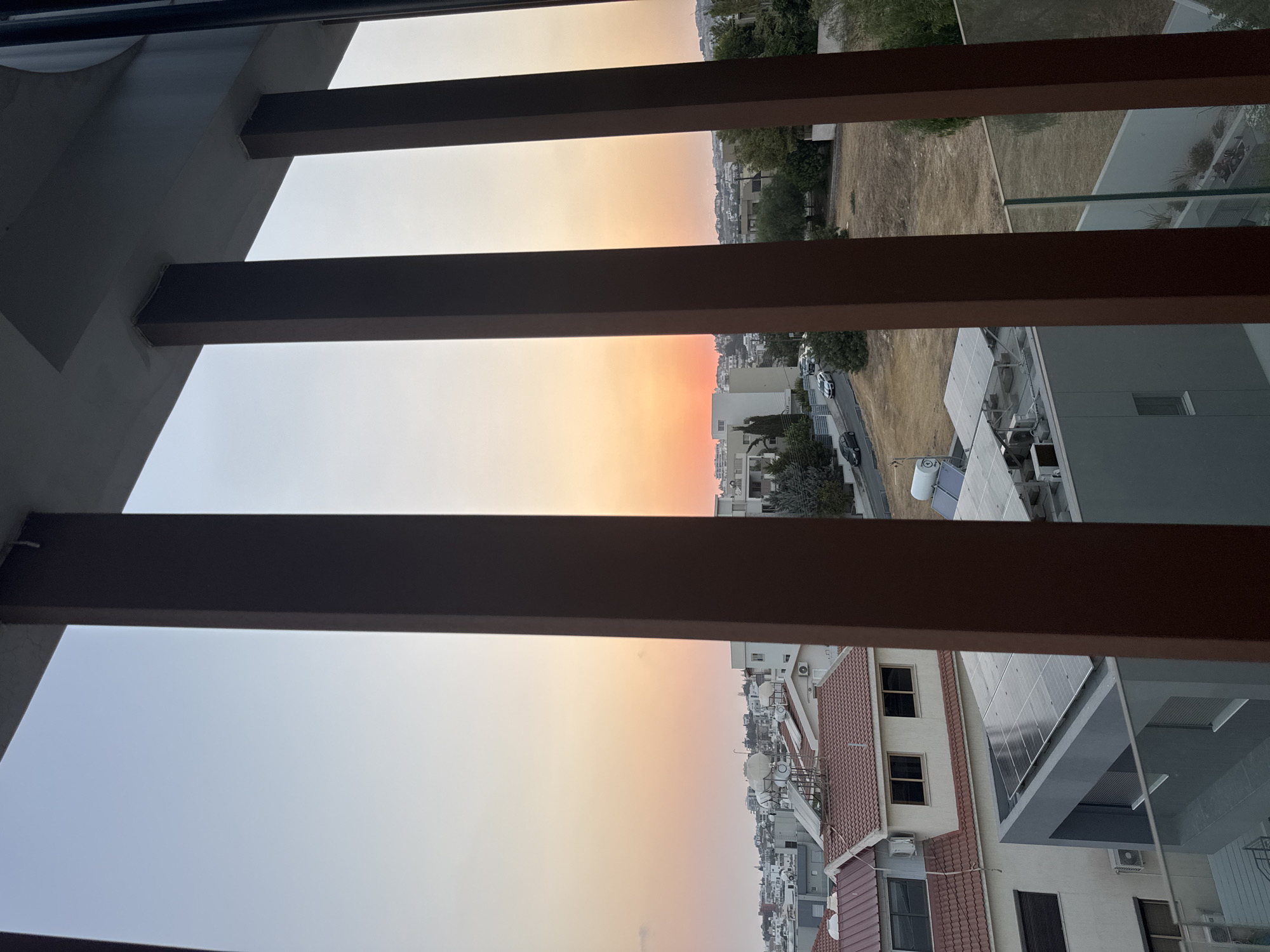 The image shows a view of Agios Athanasios Municipality, Cyprus, during sunset. The sky exhibits a gradient of colors from light blue to orange and pink, indicating the setting sun. In the foreground, there are dark brown vertical slats, part of a balcony or architectural feature, framing the scene. Beyond these slats, a residential area is visible with several buildings. Some roofs are tiled in red, while others are flat and feature solar panels. There are also visible air conditioning units on some of the buildings. A street with a few cars can be seen winding through the neighborhood. Trees and dry vegetation are scattered throughout the landscape. The overall impression is one of a quiet suburban evening in a Mediterranean city.