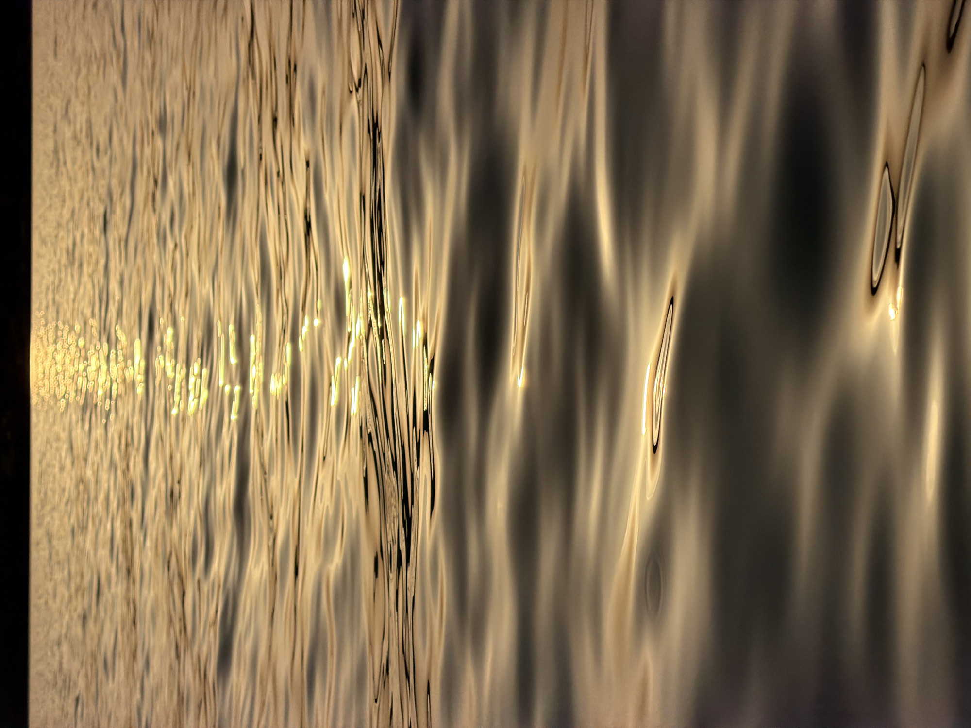 The image depicts a close-up view of rippling water with sunlight reflecting off its surface. The water is a dominant element, occupying the majority of the frame. The light creates shimmering patterns and highlights on the waves. The top of the image shows a dark, out-of-focus horizon line, possibly representing land or the sky. There are no people, animals, or man-made objects clearly visible in the scene. The overall impression is one of natural beauty and tranquility. Based on the provided information that the image is from Alghero, Italy, this scene could be of the sea or a lake near the city.