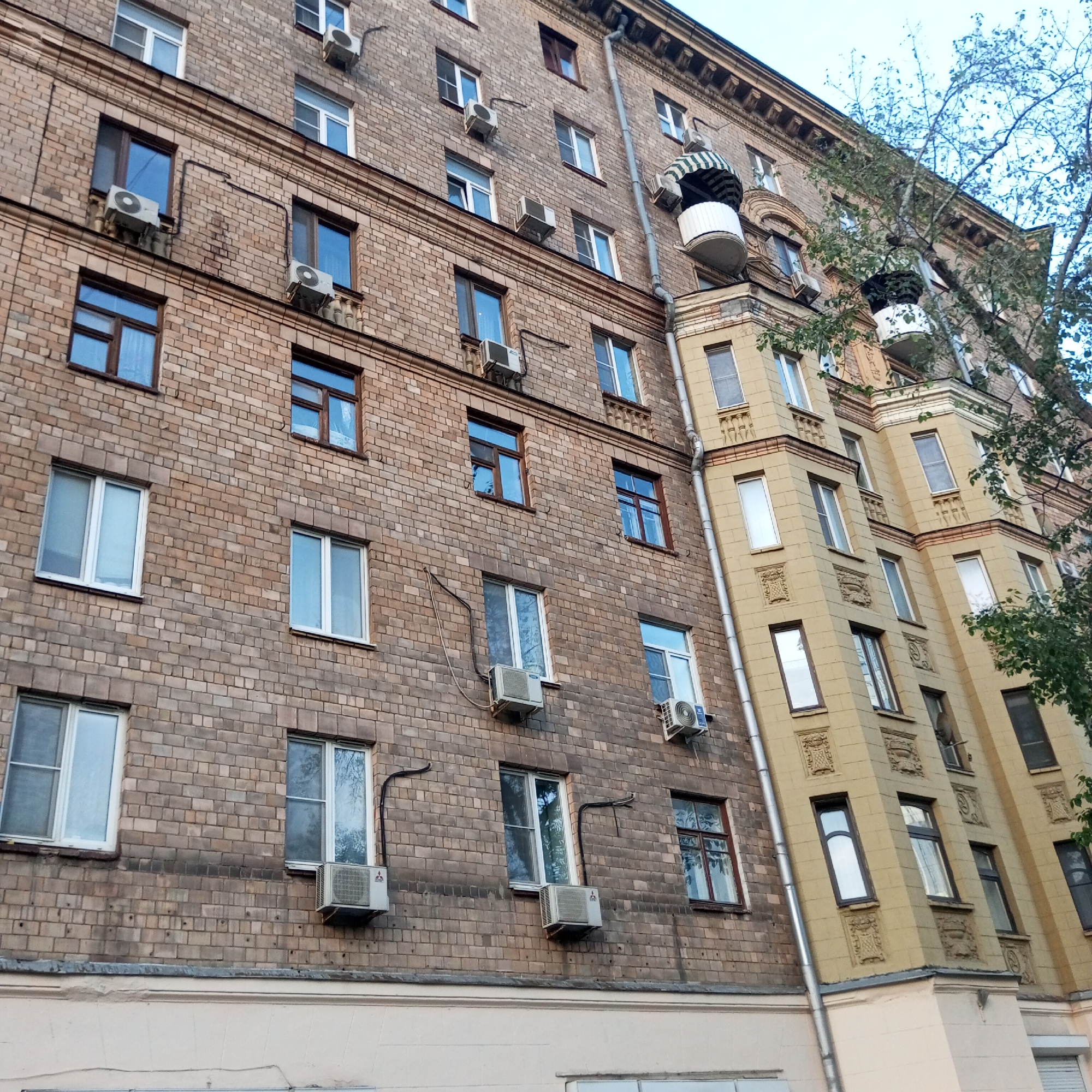 This image captures a side view of a multi-story building, likely an apartment complex, located in Moscow, Russia. The building is constructed with a combination of beige brick and lighter-colored stucco, featuring numerous windows of varying sizes, many of which are equipped with air conditioning units. The architecture suggests an older style, possibly from the Soviet era, with decorative elements on the stucco portions and a distinct balcony with an awning. A large tree with green foliage partially obscures the building on the right side of the frame. The sky above is overcast. There are no people visible in the image. The perspective is from a low angle, looking upwards at the building's facade. The overall condition of the building appears worn, with some discoloration and signs of age on the brickwork.