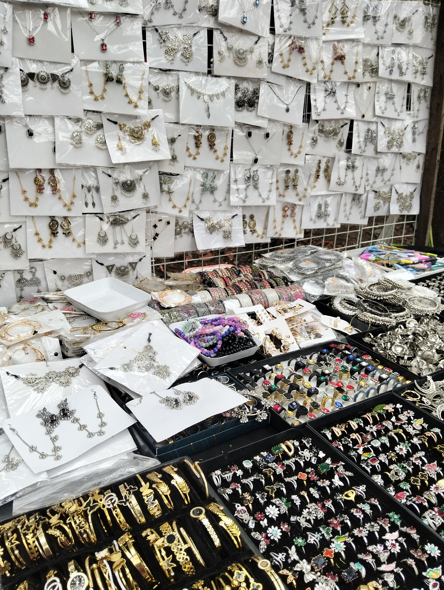 The image depicts a roadside stall in Dhaka, Bangladesh, selling a variety of costume jewelry. The stall displays a wide range of items including necklaces, earrings, bracelets, and rings. These items are arranged on a table and also hung on a mesh display behind the table. The jewelry includes pieces made from silver, gold, and colorful materials. The overall impression is one of a bustling marketplace offering affordable fashion accessories.
