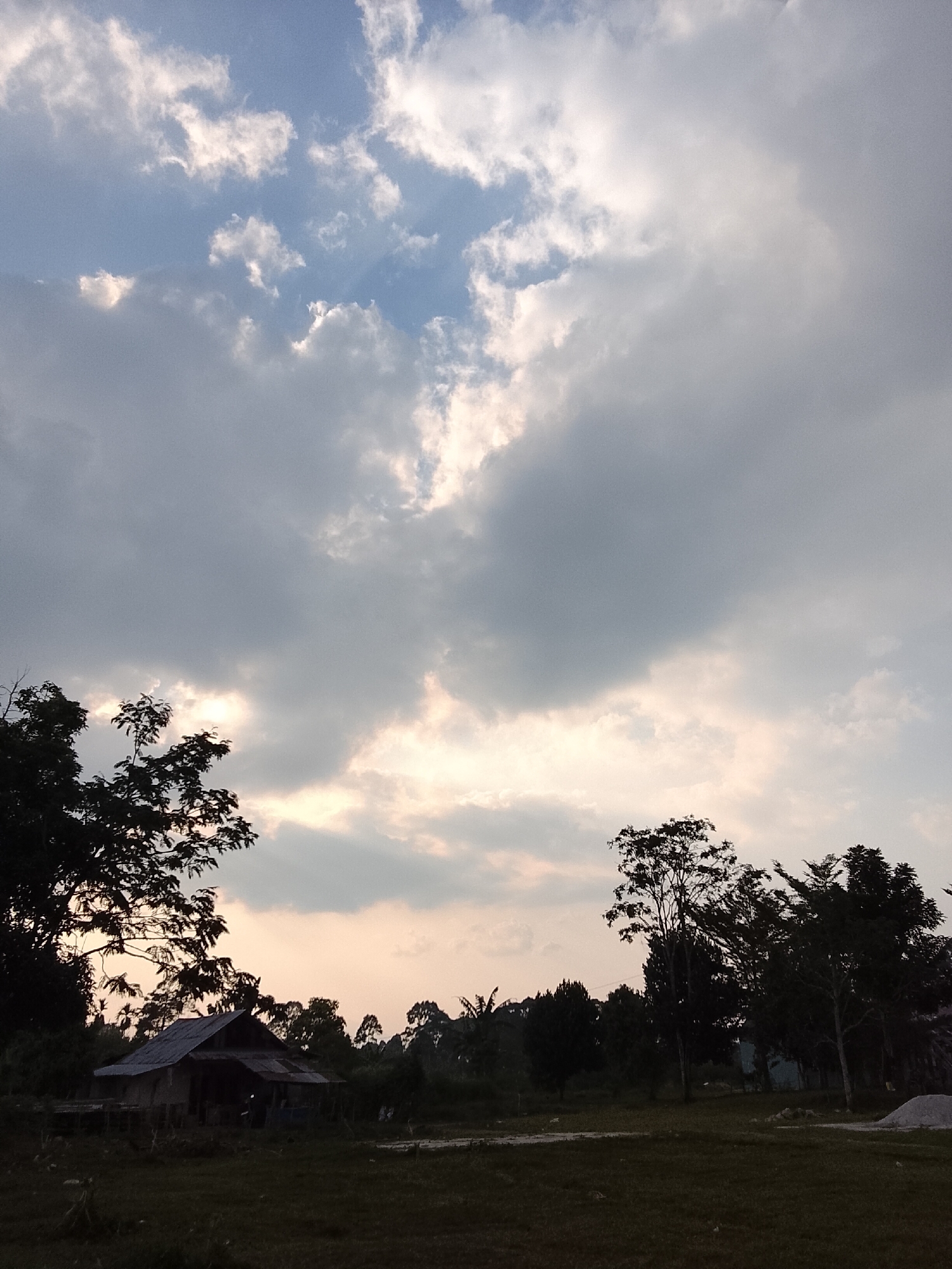 A wide, vertical shot captures a cloudy sky above a rural landscape in Padang Dalom, Indonesia. The sky is filled with large, grey and white clouds, with some patches of blue sky peeking through. Sunlight filters through the clouds, creating a soft, diffused light. In the lower third of the image, silhouetted trees frame a small, rustic house with a tin roof. The house appears to be made of wood and sits in a clearing. A dirt path is visible in front of the house, leading into a grassy field. The overall impression is one of a quiet, serene rural setting.