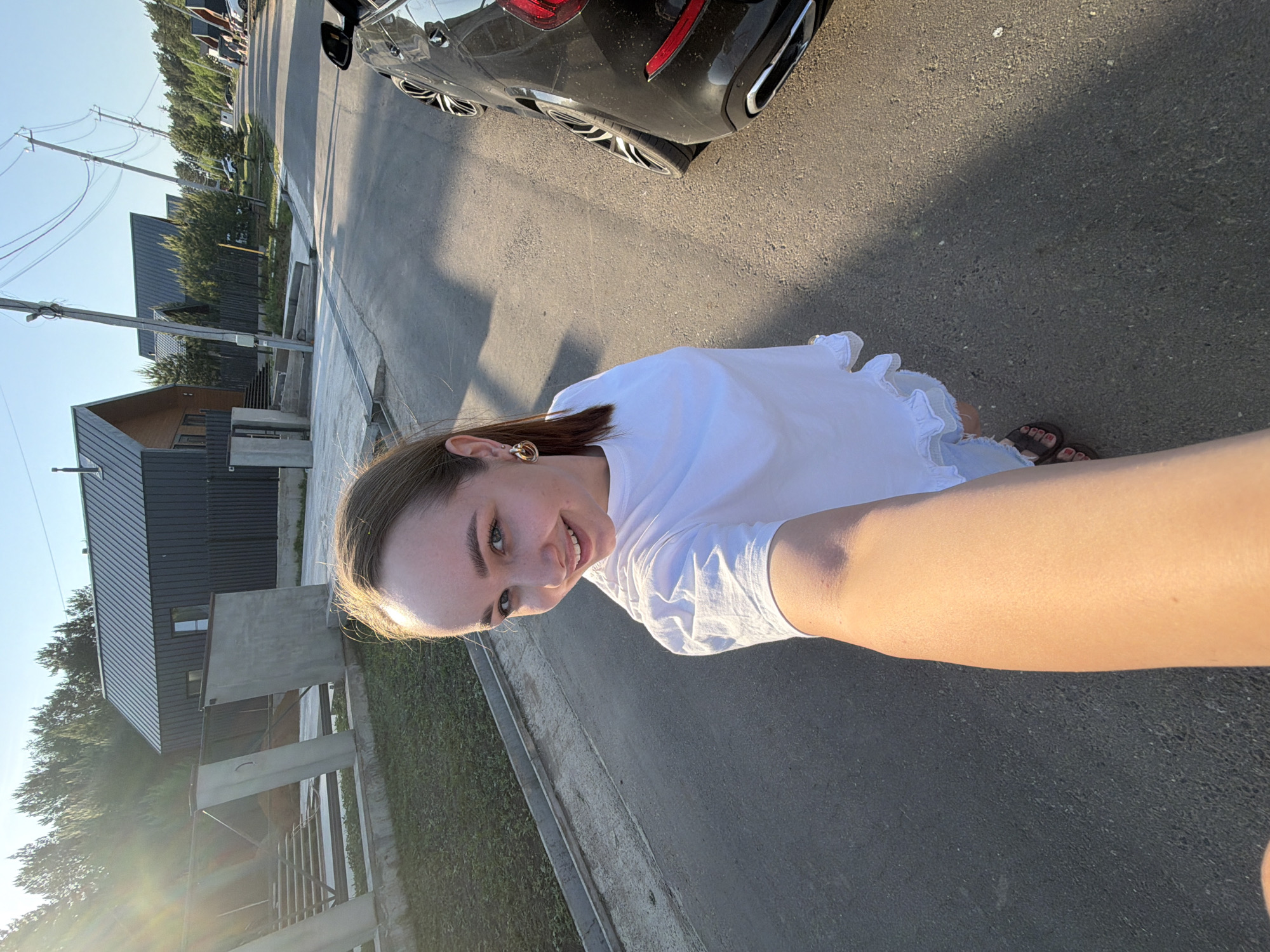 A young woman in a white t-shirt takes a selfie on a street in Kamyennoye, Russia. She's standing on the pavement near a car, with houses and greenery visible in the background. The sun is shining.