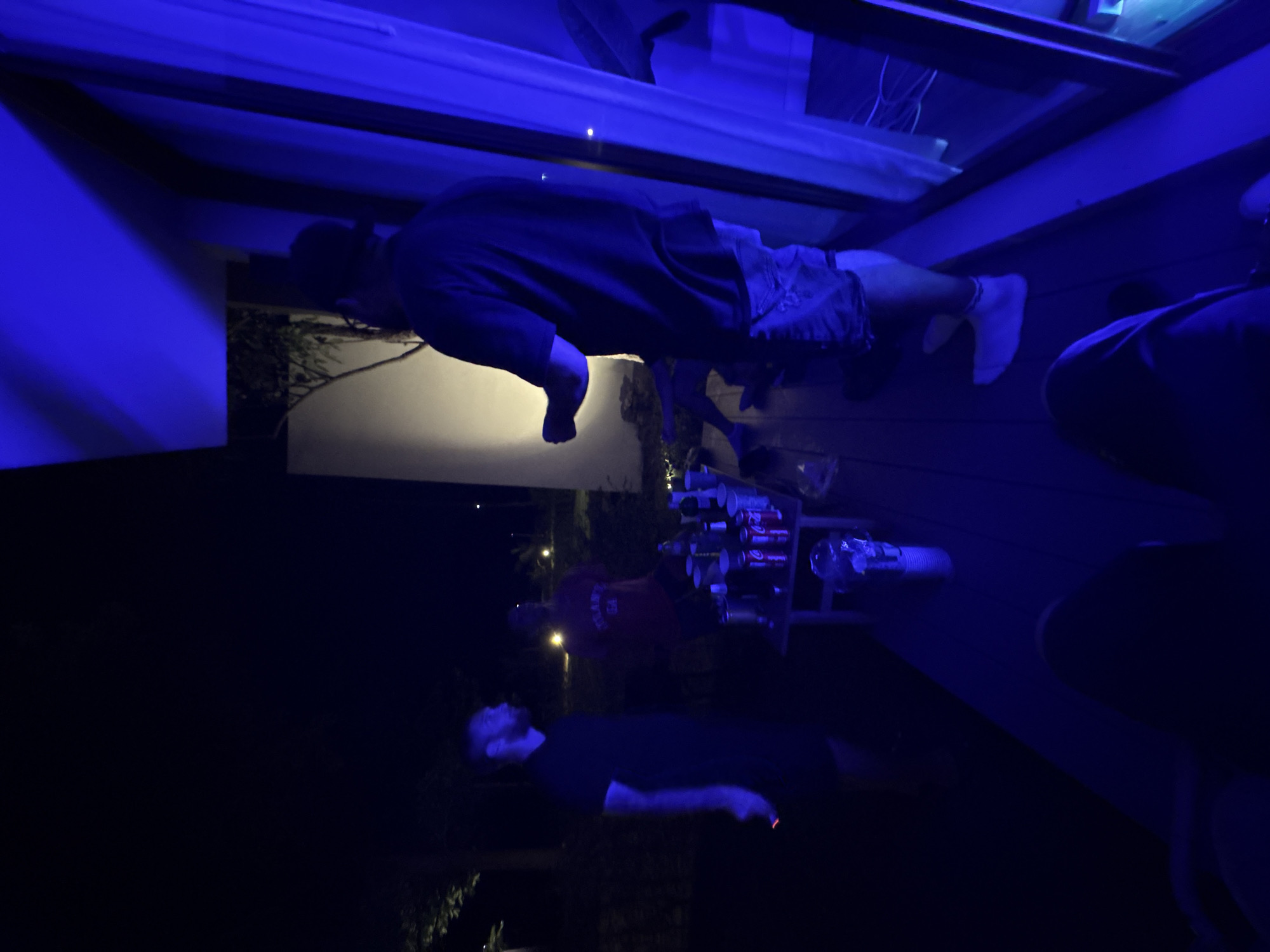 In Baan Plai Laem, Thailand, at night, four individuals are visible on a balcony. One person, wearing a dark shirt and shorts, stands with their back to the camera, illuminated by blue light. Another person, in a dark t-shirt, stands to their left, facing away from the camera with a lit cigarette in hand. A third person, wearing a red shirt, is partially visible behind a table laden with drinks and cups. The fourth person's lower body is visible in the foreground, with their legs crossed. The balcony is furnished with a small table and chairs. The scene is lit by artificial blue lights, creating a dark and somewhat blurred atmosphere.