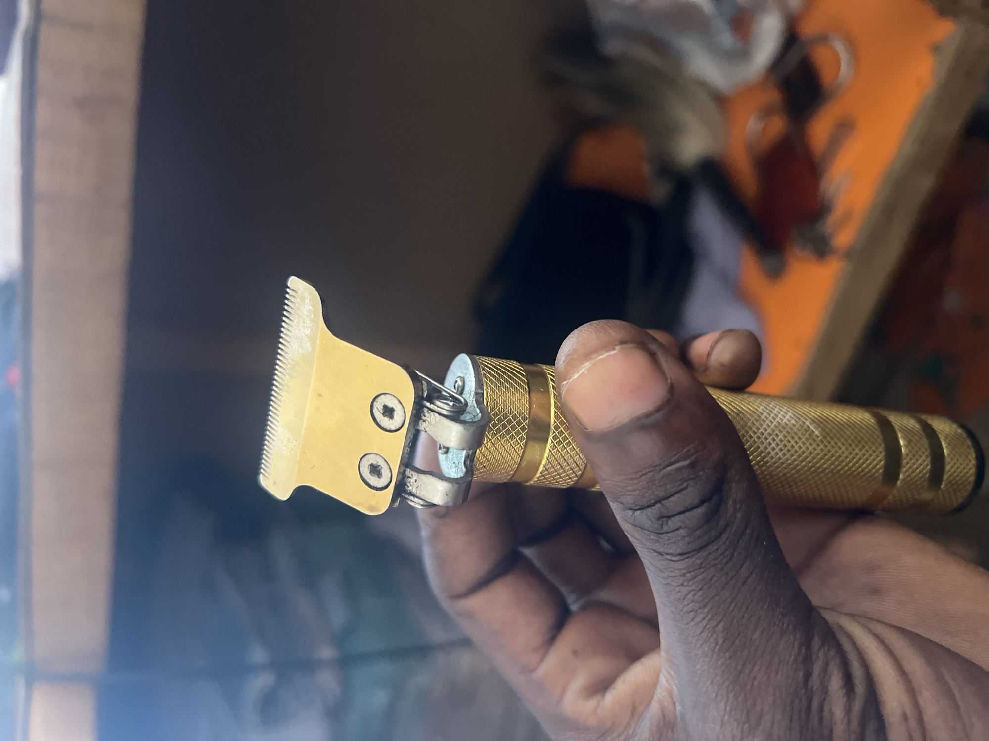 The image shows a close-up of a gold-colored electric trimmer being held by a dark-skinned hand. The trimmer has a golden blade with visible teeth and two small screws holding it in place. The handle of the trimmer is also gold and has a textured, diamond-like pattern. The background is out of focus, but appears to be an indoor setting with shelves and some colorful objects, possibly on a desk or workbench. The lighting suggests it is daytime. The image's origin within Jalingo, Nigeria, is unknown.