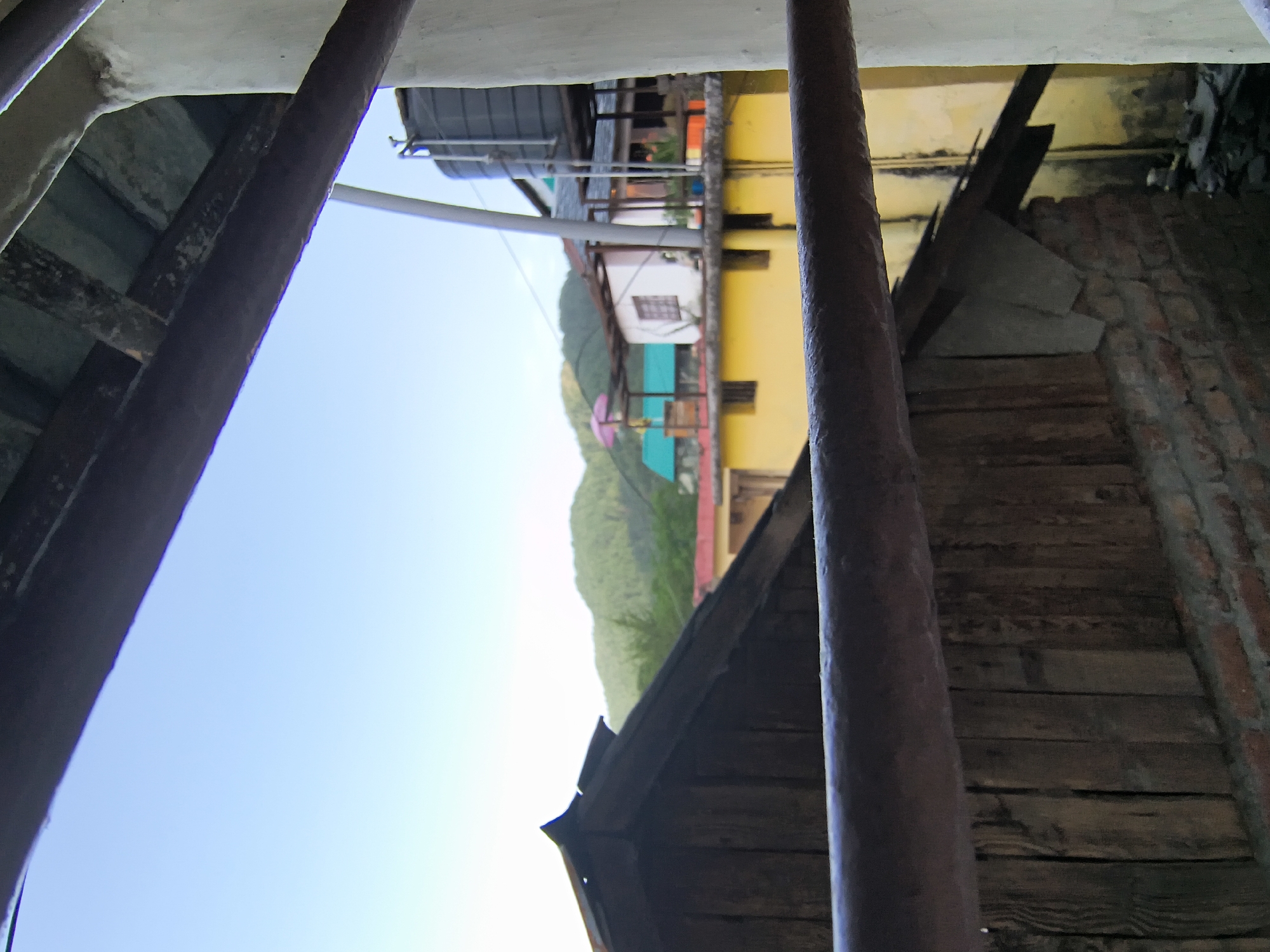 The image depicts a view from behind metal bars, potentially a window or enclosure, in Haini, India. The foreground consists of the bars, weathered wood, and a partial brick wall. Through the bars, the background reveals a scene of buildings with yellow, teal, and white facades, set against a backdrop of a tree-covered mountain and a clear blue sky. A utility pole is also visible.