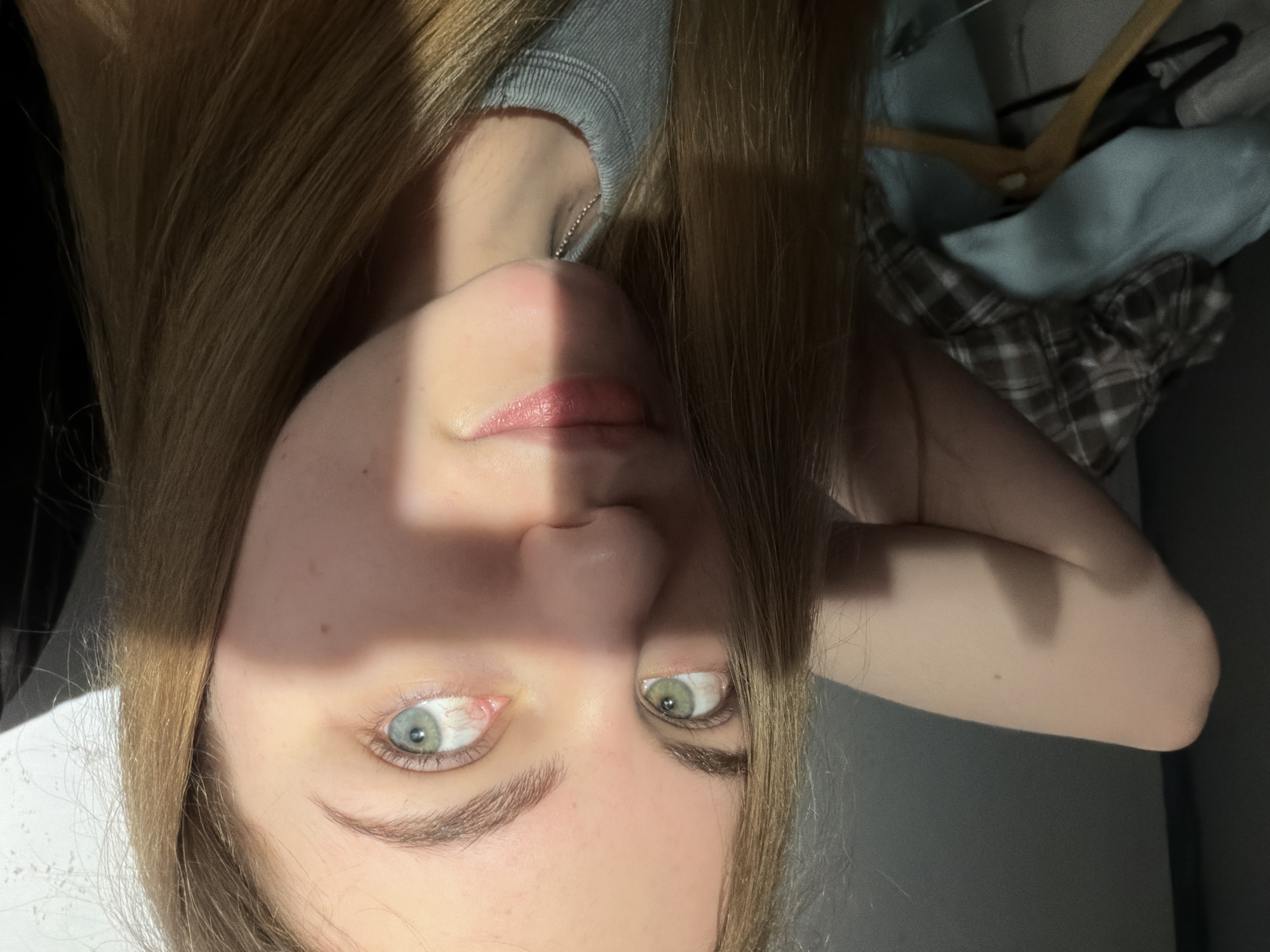 A young woman with long, blonde hair is captured in a close-up, head-and-shoulders portrait. She is facing the camera directly, with her left arm bent and resting behind her head. Her eyes are a striking heterochromatic blue-green, with her left eye being a lighter shade of green and her right eye a pale blue. A harsh shadow diagonally crosses her face, obscuring the left side of her nose and mouth, and a portion of her left eye. Her lips are slightly parted, and she appears to be wearing a light blue, ribbed-top garment. In the background, to the left, a pile of folded clothes, including a plaid shirt and a light blue garment, is visible, along with a wooden hanger. The lighting suggests an indoor setting, possibly near a window, given the strong directional shadow. The overall environment is dimly lit, with the exception of the area illuminated by the light source.