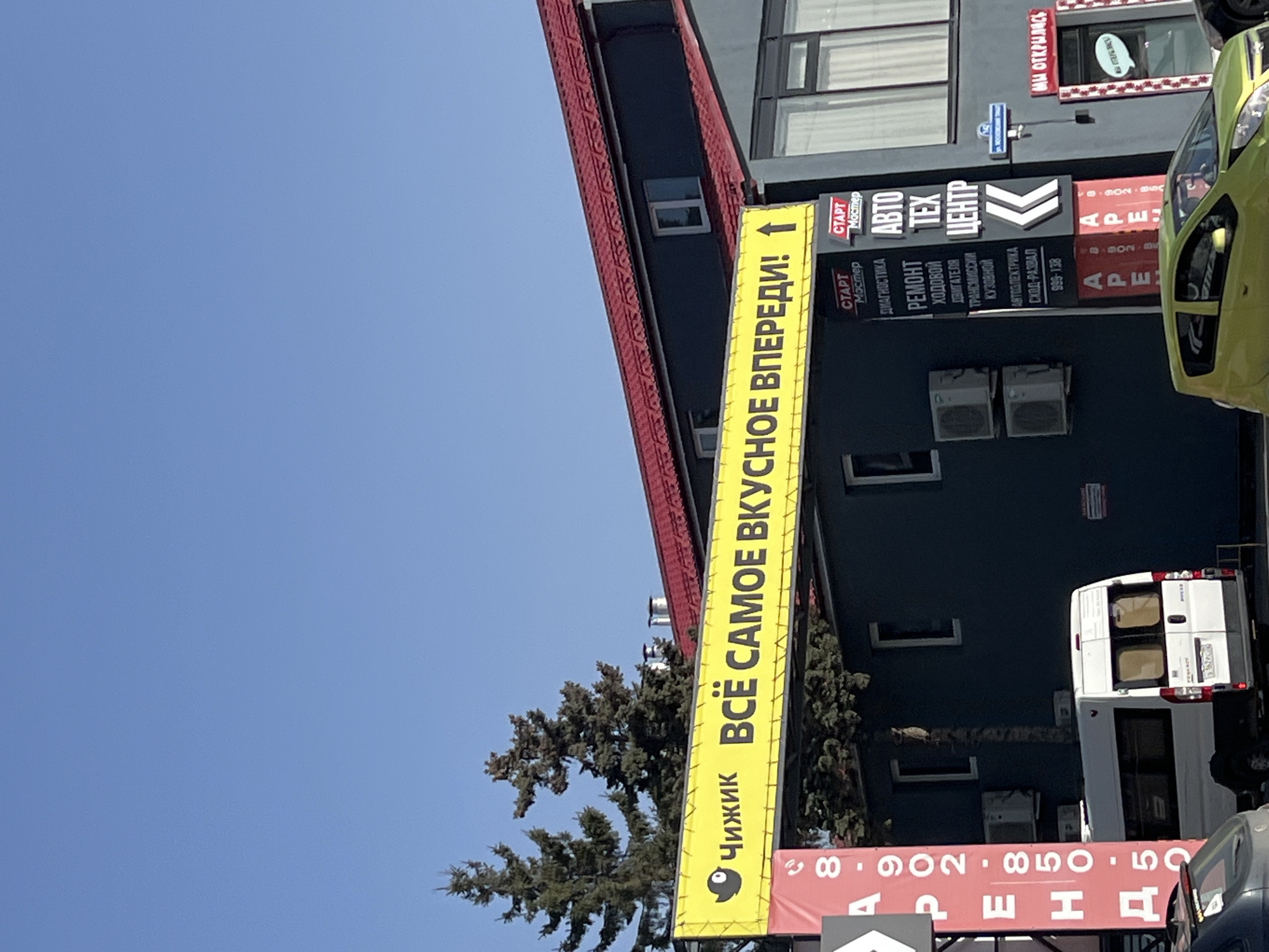 The scene depicts an outdoor urban environment under a clear blue sky, located at Novaya Zarya Street 27 in Tyumen, Russia. A large yellow banner, featuring a black bird logo and the Russian text "ЧИЖИК ВСЁ САМОЕ ВКУСНОЕ ВПЕРЕДИ!" (CHIZHIK ALL THE TASTIEST IS AHEAD!), spans horizontally across the upper middle section of the image, above a two-story building. The building has dark grey walls and a red corrugated metal roof, with multiple rectangular windows and white external air conditioning units mounted on its facade. To the right of the yellow banner, a tall, black and red sign advertises "СТАРТ Мастер АВТО ТЕХ ЦЕНТР" (START Master AUTO TECH CENTER) and lists services including diagnostics, repair of chassis, engine, transmission, bodywork, auto electrics, and wheel alignment, along with a phone number (995-138). Below this sign, two vertical red banners display "АРЕНДА" (FOR RENT) with phone numbers. A blue street sign indicating "ул. Новая Заря 27" is affixed to the building's exterior. A red-framed doorway on the far right displays "МЫ ОТКРЫЛИСЬ" (WE ARE OPENED). In the foreground, a white cargo van, possibly a Peugeot Boxer (partial license plate "У426РТ 152"), is parked or moving left below the yellow banner. A lime green compact car is partially visible in the lower right, and the rear of a dark-colored vehicle is in the lower left. Several coniferous trees are visible behind and to the left of the building. No individuals are present in the frame.