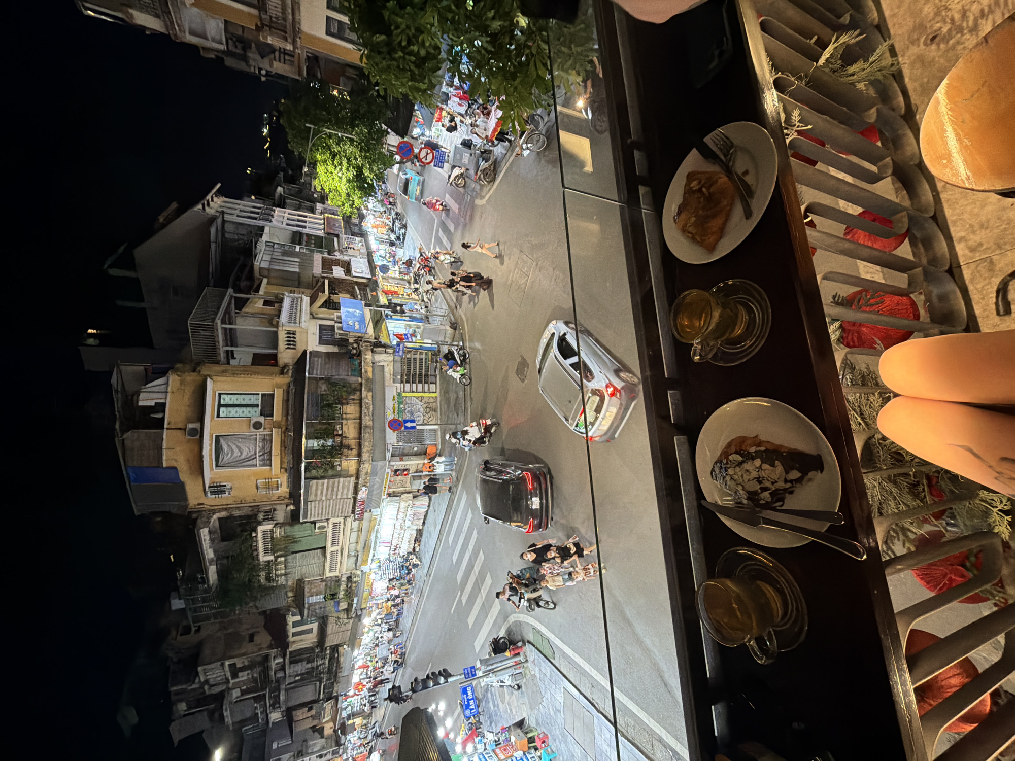 The image presents a vibrant nighttime street scene in Hà Nội, Vietnam, viewed from an elevated perspective, likely a cafe balcony or window. In the immediate foreground, a dark wooden table or railing holds two white plates with desserts – one partially eaten pastry and another with a dessert topped with chocolate and slivered almonds. Two clear glass cups of tea rest on saucers beside the plates. Part of a person's legs and knees are visible to the right, suggesting a seated individual. The balcony railing features decorative elements like red, textured ornaments and greenery.
The midground reveals a bustling street illuminated by streetlights, vehicle lights, and the glow from numerous shopfronts. The road is busy with many motorbikes and a few cars. A distinct crosswalk is visible, along with traffic signals. People are actively moving, including pedestrians on sidewalks and a group, possibly a family with children, gathered near a scooter in the middle of the road.
The background consists of densely packed, multi-story buildings lining both sides of the street. These structures display varied architecture, some with balconies and air conditioning units. Trees are interspersed among the buildings, adding greenery to the urban environment. The sky is dark, confirming the nighttime setting. A blue street sign is partially visible, reading "LAN KINH," likely part of a street name such as Lãn Ông. The overall scene conveys a lively and energetic urban atmosphere after dark.