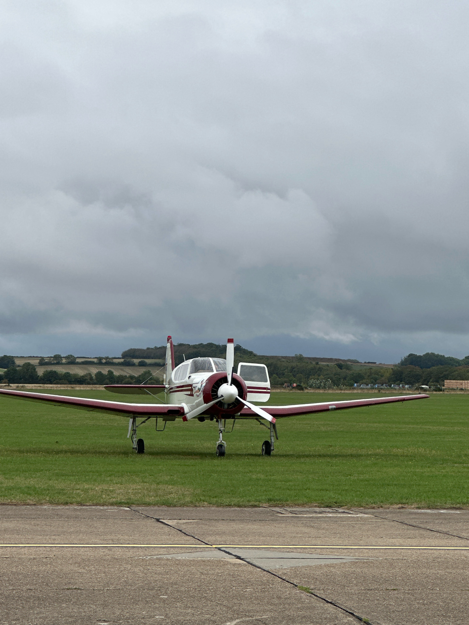 A small, twin-engine propeller airplane, predominantly white with maroon trim, is parked on a grassy airfield under a cloudy sky. The airplane's door is open, and the propeller is stationary. The airfield is bordered by a concrete taxiway in the foreground. In the background, rolling hills with trees and fields are visible. The image appears to be taken outdoors on a somewhat overcast day. No people are visible in the image. The location is identified as Cambridge, United Kingdom.