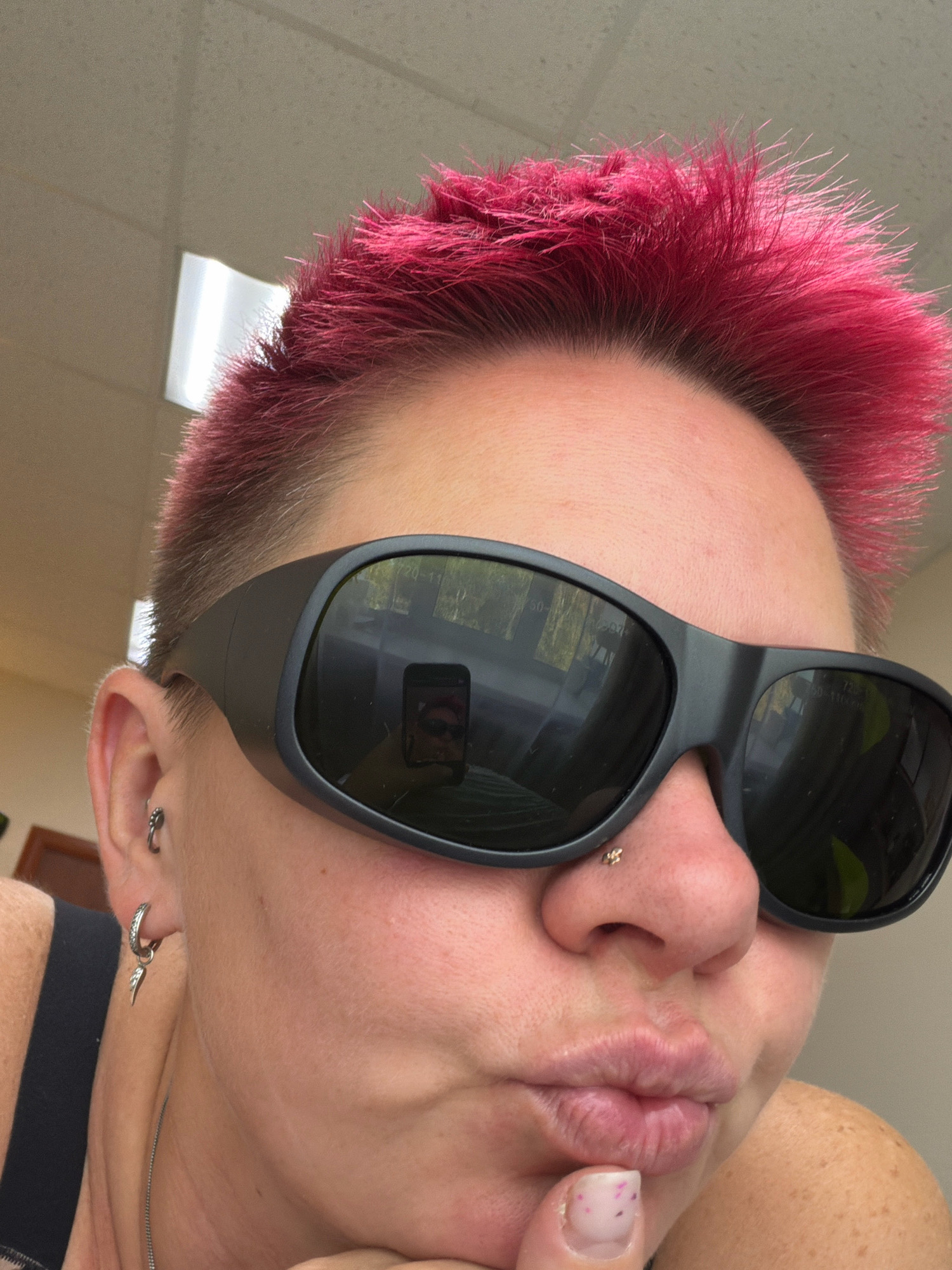 A close-up selfie shows a person with short, spiky, bright pink hair. They are wearing large, black wrap-around sunglasses, which reflect a handheld mobile phone in use. The person has a nose piercing and two earrings: one on the helix of their left ear and another dangly earring with a wing charm on their earlobe. Their lips are pursed as they hold a finger to their chin. The background appears to be an indoor environment with a tiled ceiling and fluorescent lighting. There is no discernible location in Chaikovsky, Russia, or any other city.