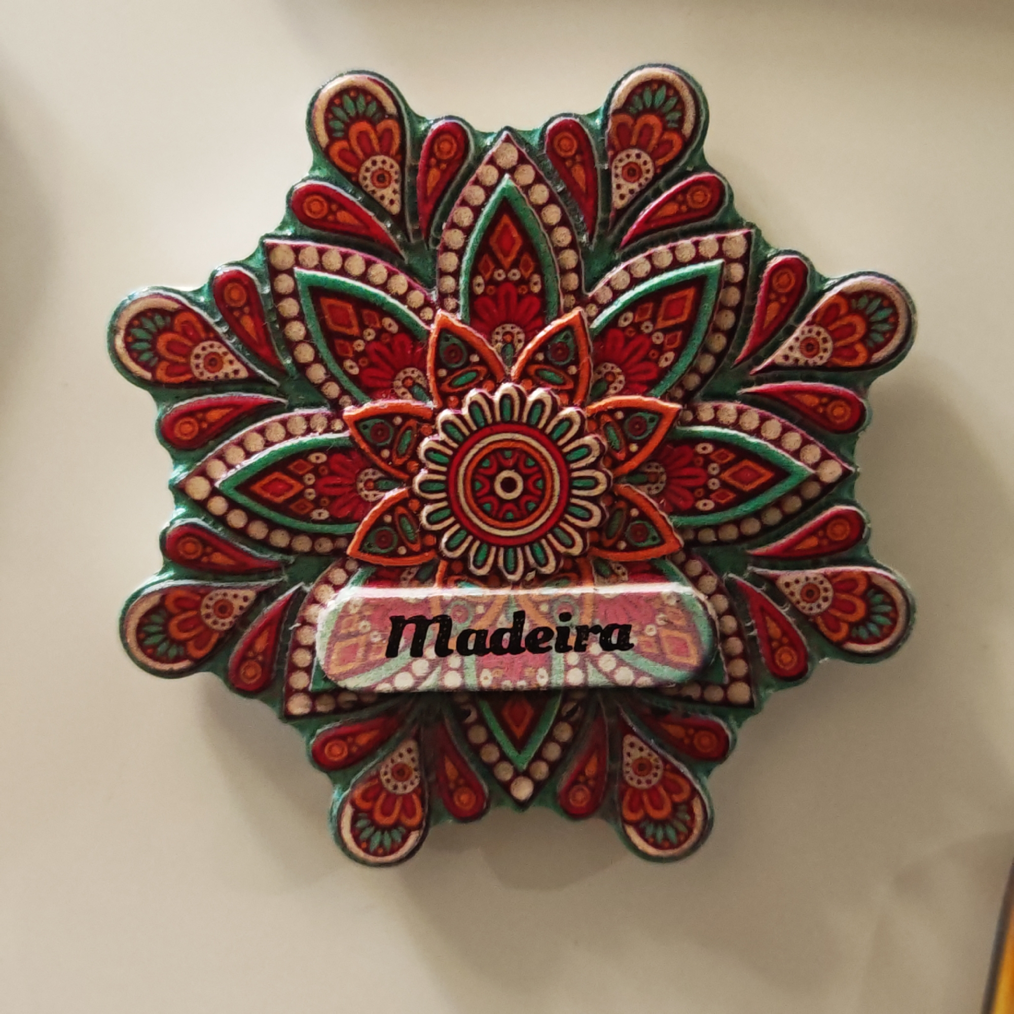 This media file captures a close-up of a vibrantly colored, mandala-style decorative object, possibly a souvenir magnet, set against a plain, light-colored background. The object features an intricate, symmetrical design with an octagonal shape and petal-like extensions around its perimeter.
The dominant colors are rich reds, oranges, and greens, interspersed with numerous small white or cream dots that outline various patterns and shapes, giving the object a textured, possibly hand-painted, appearance. A prominent floral motif with concentric rings is at the center.
Across the lower-middle section of the decoration, a rectangular panel displays the word "Madeira" in stylized black text on a light pinkish background that has a faint geometric pattern. The item appears to be static, with no people or activity shown. The lighting is even and bright, highlighting the details and colors of the object. While the object itself names "Madeira," the provided context places this media file in Novo-Nikol'skoe, Russia.