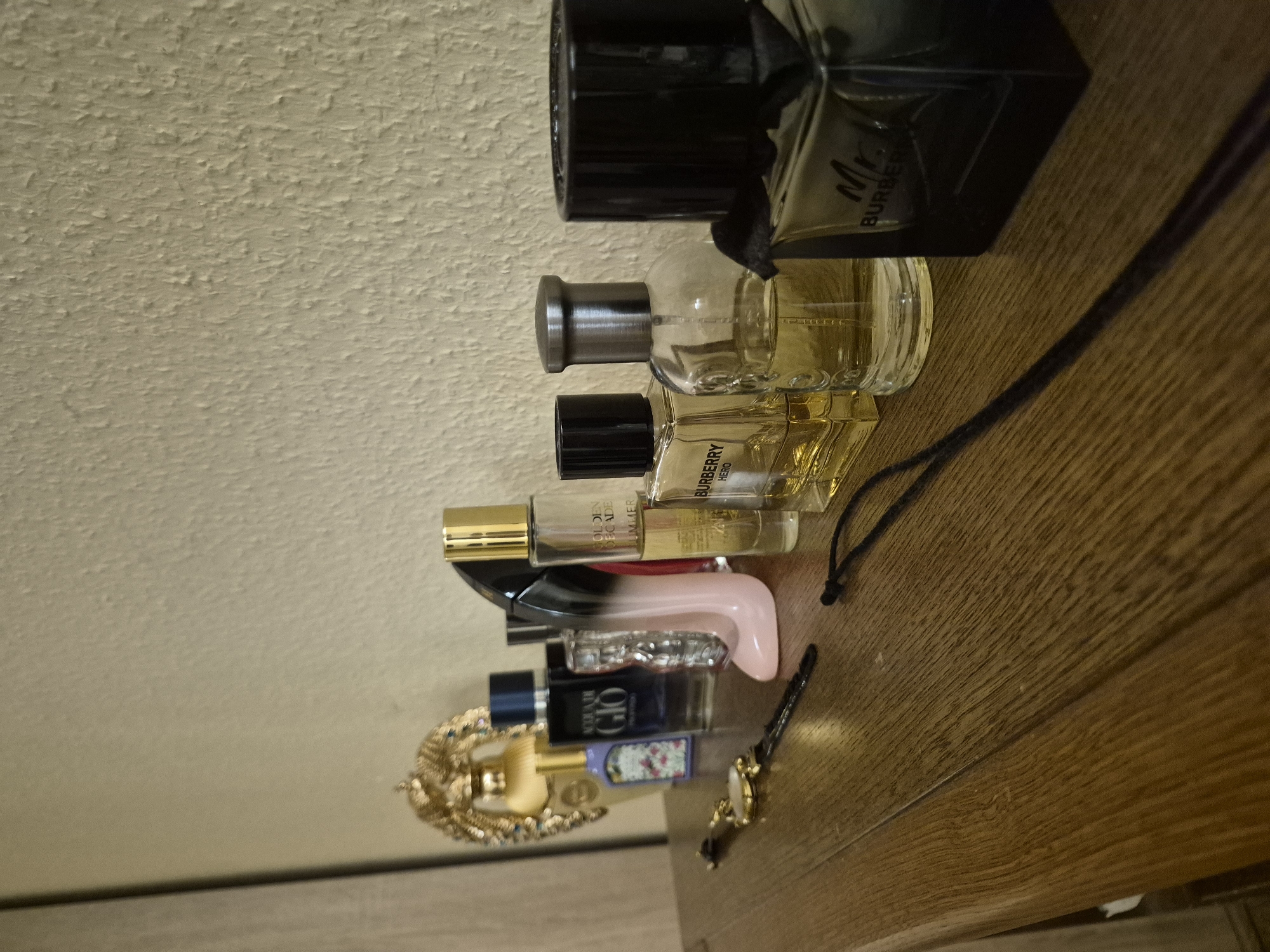 The image displays a collection of perfume bottles arranged on a wooden surface, with a textured, off-white wall in the background. The bottles vary in size, shape, and color, with several appearing to be clear glass filled with golden or light-colored liquid, and at least one being a dark, opaque bottle. Some bottles have metallic caps and accents. One bottle clearly visible has the text "BURBERRY HERO" on it. Another bottle, partially visible at the top, has "Mr. Burberry" written on it.
Other objects on the wooden surface include a gold-colored bracelet or necklace with intricate detailing and a wristwatch with a gold-colored face and black band. A dark cord, possibly from a tag or another accessory, snakes across the wooden surface. The lighting suggests an indoor setting, possibly a shelf or dresser, with soft shadows cast on the wood. The overall impression is that of a personal collection of fragrances and accessories.