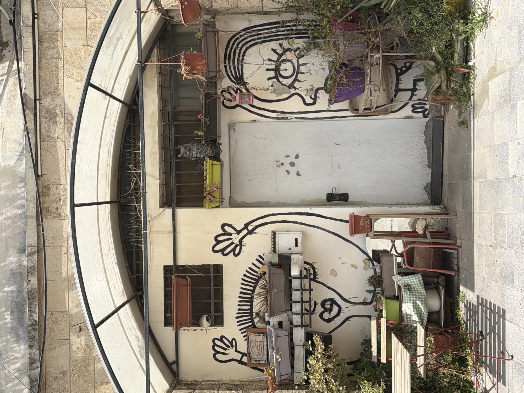 The image, taken from an elevated perspective, depicts an eclectic and artistic outdoor space, likely an alleyway or a small courtyard in Tel Aviv, Israel. The central feature is a light-colored wall adorned with bold black graffiti, depicting stylized, whimsical figures. One prominent figure, spanning the upper half of the wall, features a large, open eye with radial lines and what appears to be a head with flowing lines. Below this, a plain white door is visible, and further down the wall, another graffiti figure shows two hands reaching out and a heart-like shape.
The area is furnished with a mix of outdoor items, creating an informal and lived-in atmosphere. Various chairs, including red metal ones, a purple fabric chair, and a wicker-style chair, are scattered or stacked to the right of the wall. Wooden tables and numerous planters, some containing lush green foliage, are placed around the space. Potted plants hang from above and rest on ledges, contributing to the verdant feel. An owl figurine is visible on a ledge near a window protected by metal bars, while another window lower down the wall features a reddish shutter.
Overhead, a translucent arched canopy structure with a dark frame provides partial shelter. The ground is paved with light-colored stones, and sharp shadows are cast, indicating a bright and sunny daytime setting. The scene suggests a bohemian or artistic establishment or private area. No discernible text is visible within the image.