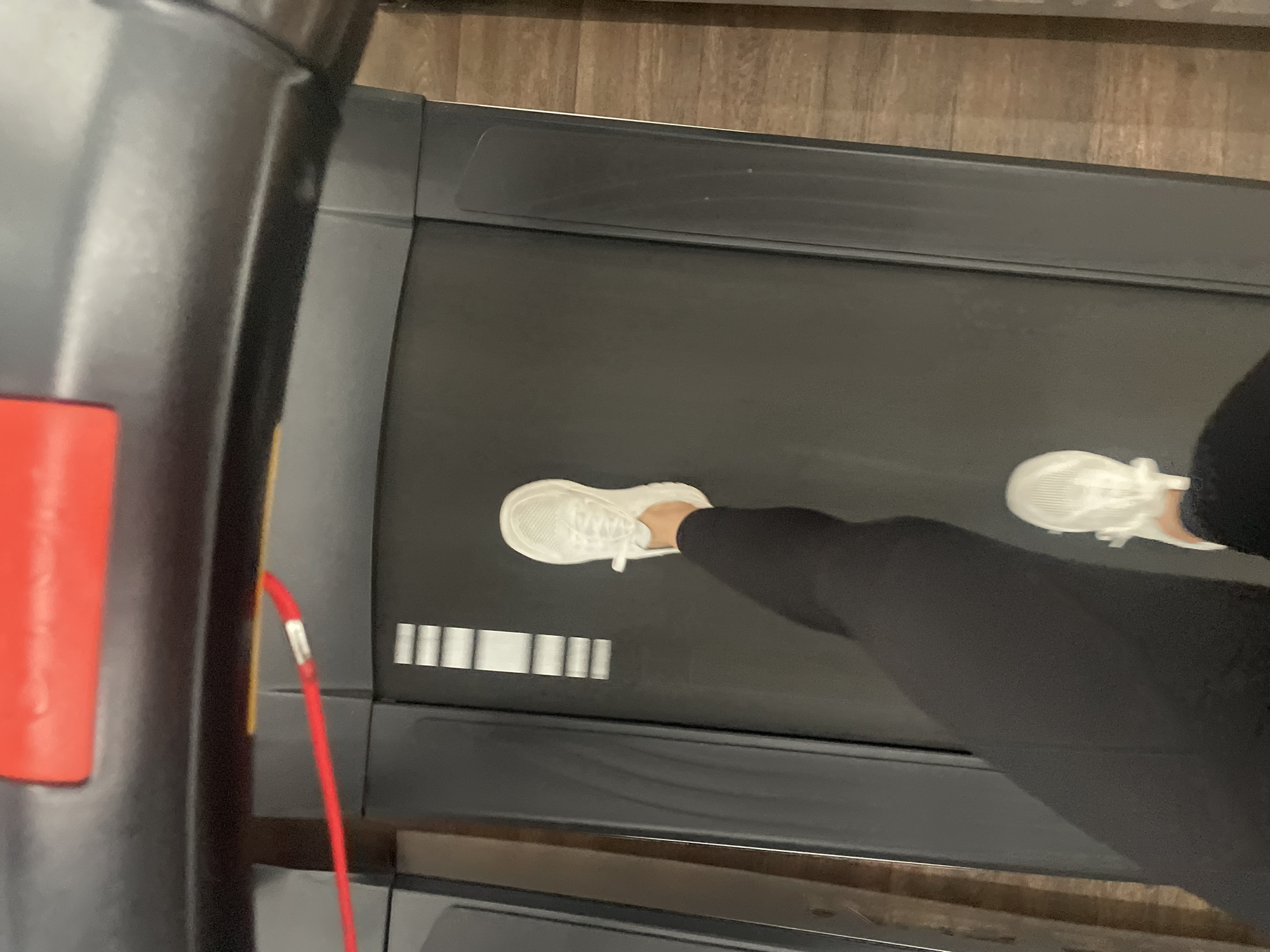 An overhead, slightly angled shot captures a person's legs and feet actively using a treadmill. The person is wearing dark athletic leggings and white athletic shoes. One foot is shown planted on the dark gray treadmill belt, while the other is partially visible near the top right, suggesting movement or mid-stride.
The treadmill belt features several white rectangular markings that appear to be part of its design. On the left side of the image, part of the treadmill's frame is visible, along with a prominent red emergency stop clip attached to a red cord. The background reveals a light brown floor with a wood-grain pattern, indicating an indoor setting, likely a gym or home gym. The lighting is uniform, and there are no discernible details regarding the time of day, weather, or the person's emotions. No legible text is visible in the image. The activity depicted is exercising on a treadmill in an indoor facility in Sofia, Bulgaria.
