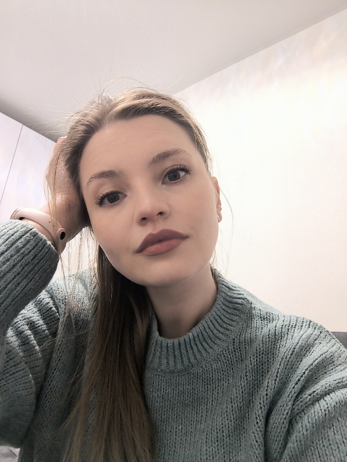 A young woman with light skin and blonde hair looks directly at the camera. She wears a textured, light blue-green sweater. Her makeup is subtle with a matte lip color. Her left hand is resting on her head. A peach-colored smartwatch is visible on her left wrist. The background includes a white wall and part of a white cabinet. The lighting is soft and even.