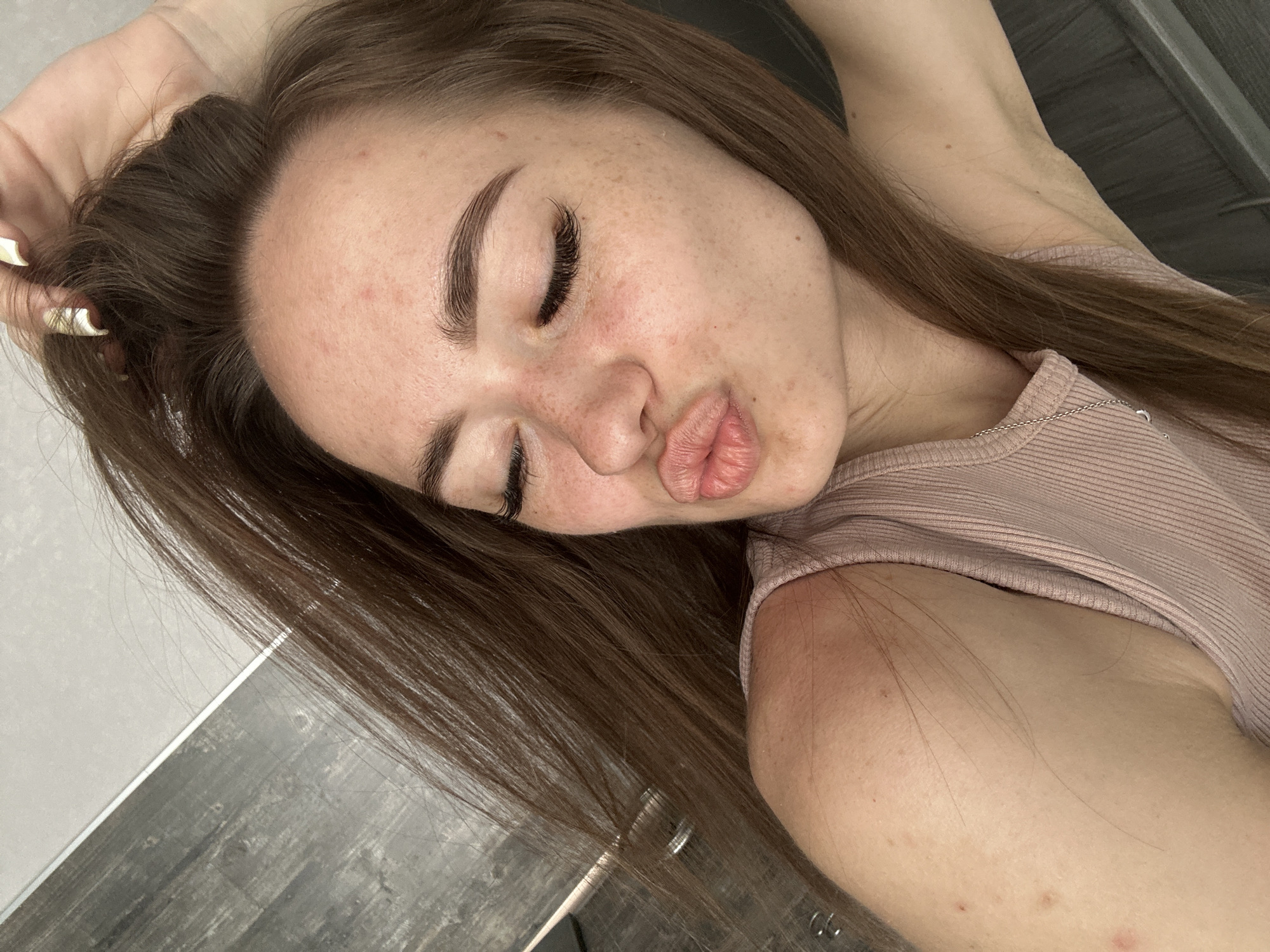 A woman with brown hair is lying down with her head on her arm and her eyes closed. She has on a pink ribbed tank top. Her lips are puckered as if she is about to kiss someone or something. The background appears to be a tiled floor.