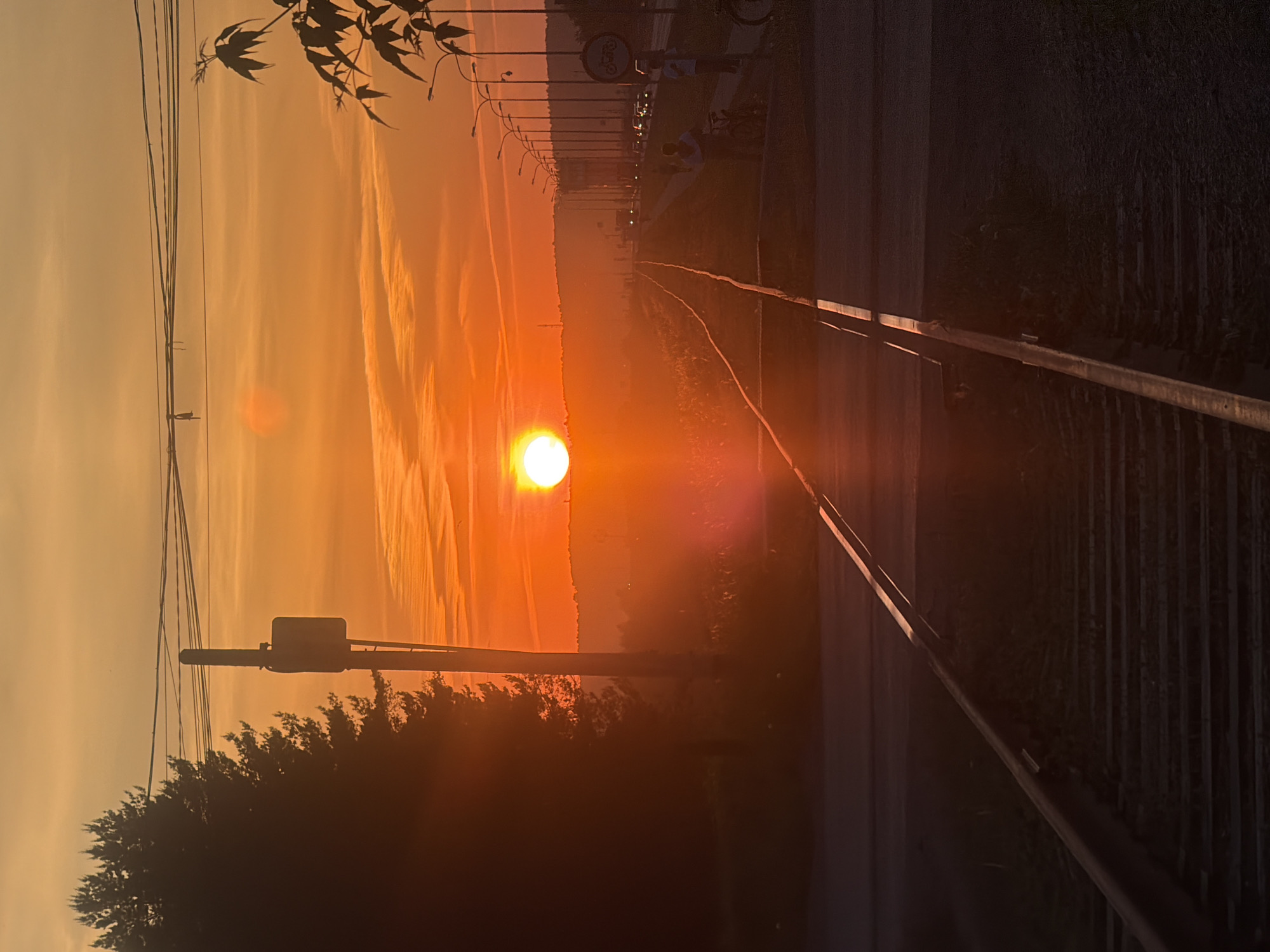 The image depicts a sunset along railroad tracks in what is probably Chaikovsky, Russia. The sun is low on the horizon, visible between the converging rails, creating a strong glare. The sky is layered with orange and yellow hues, with light cloud patterns. Utility poles with wires and streetlights line the right side of the tracks. Vegetation, possibly trees, is visible along the edges. A bike path is to the right of the rails, with people standing/sitting. The overall scene evokes a sense of tranquility and a long commute.